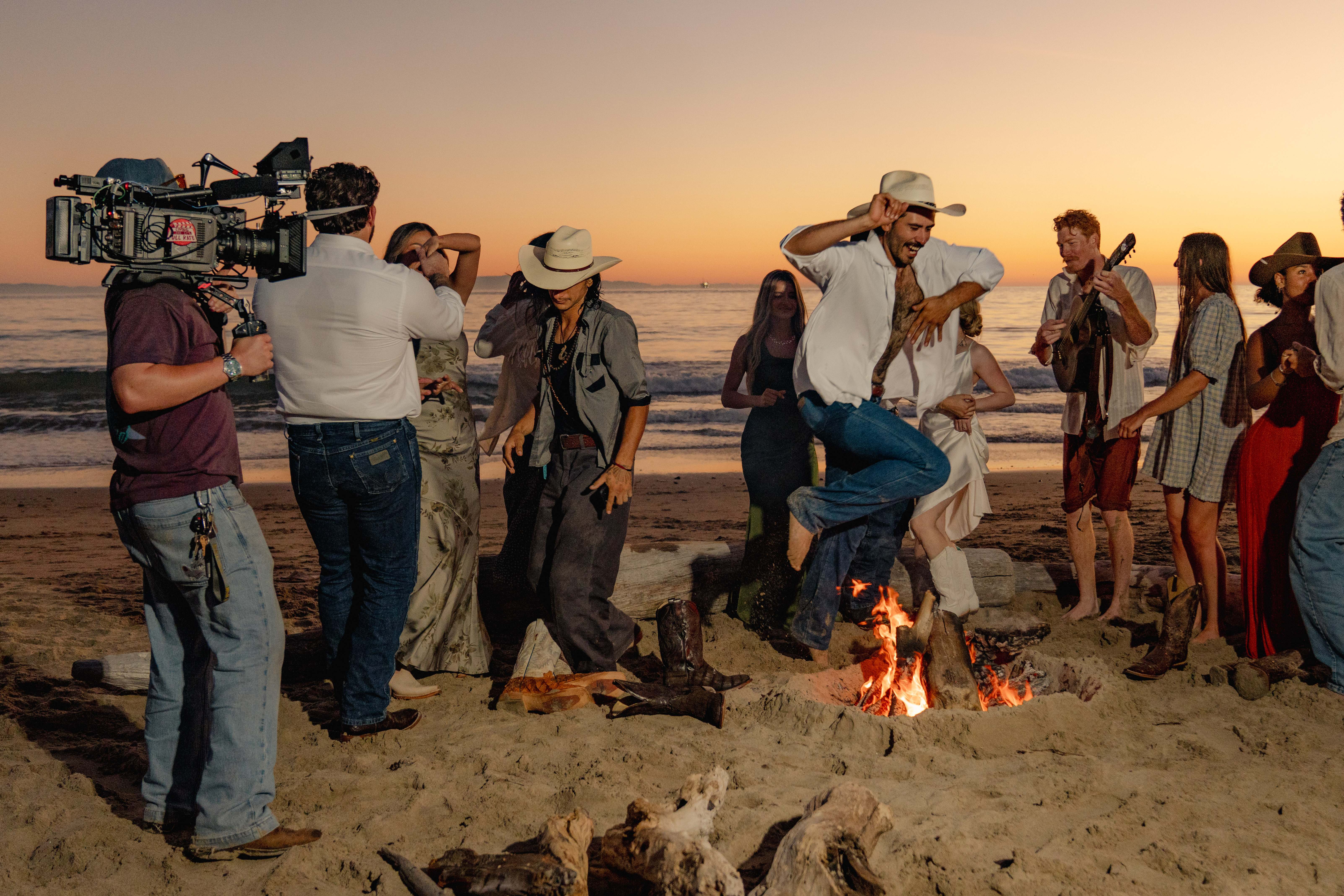 Behind the scenes: a cinematographer films guests dancing around a beach bonfire at sunset