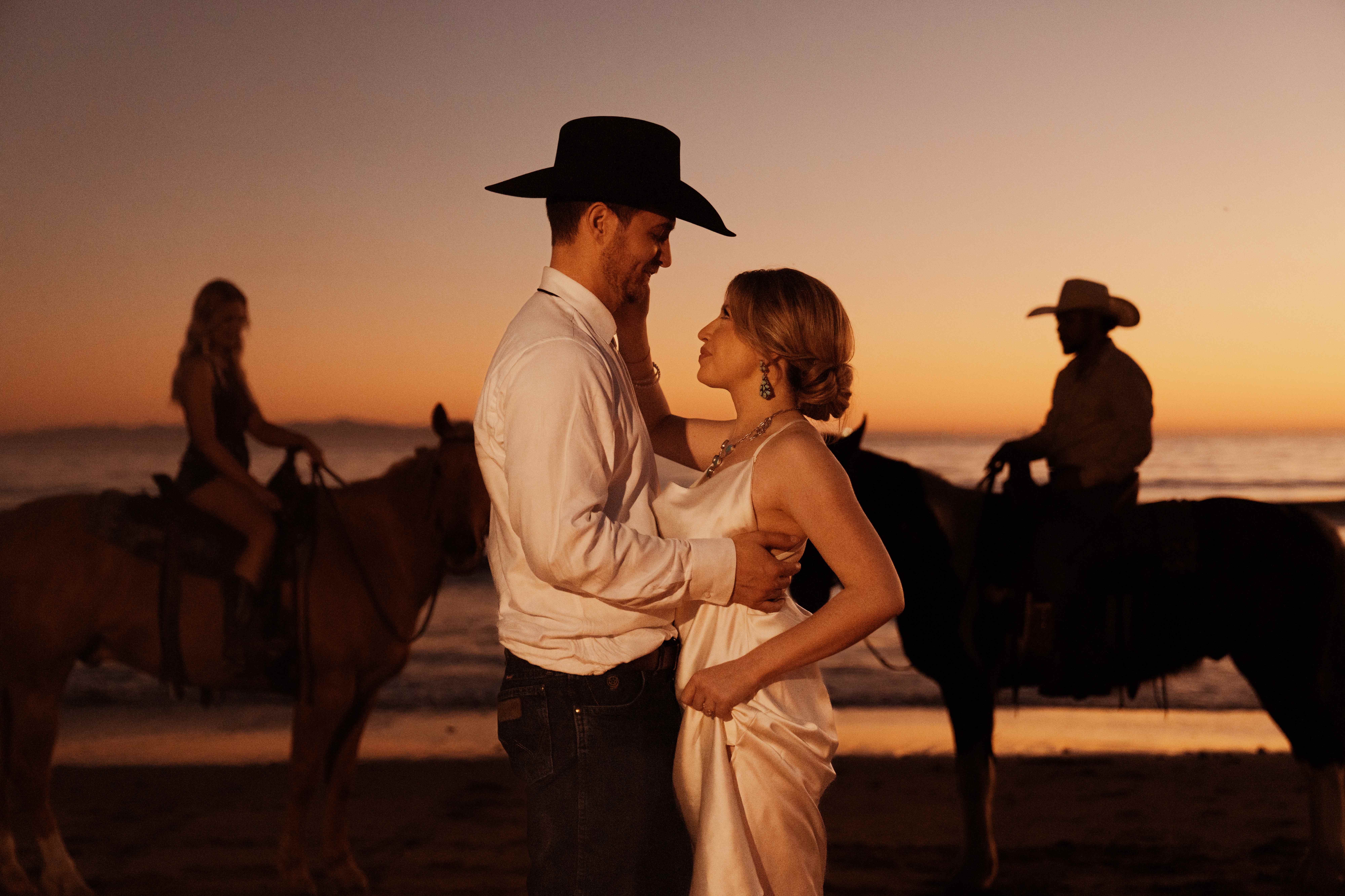 Couple embracing at golden hour on the beach with two horseback riders silhouetted against the setting sun