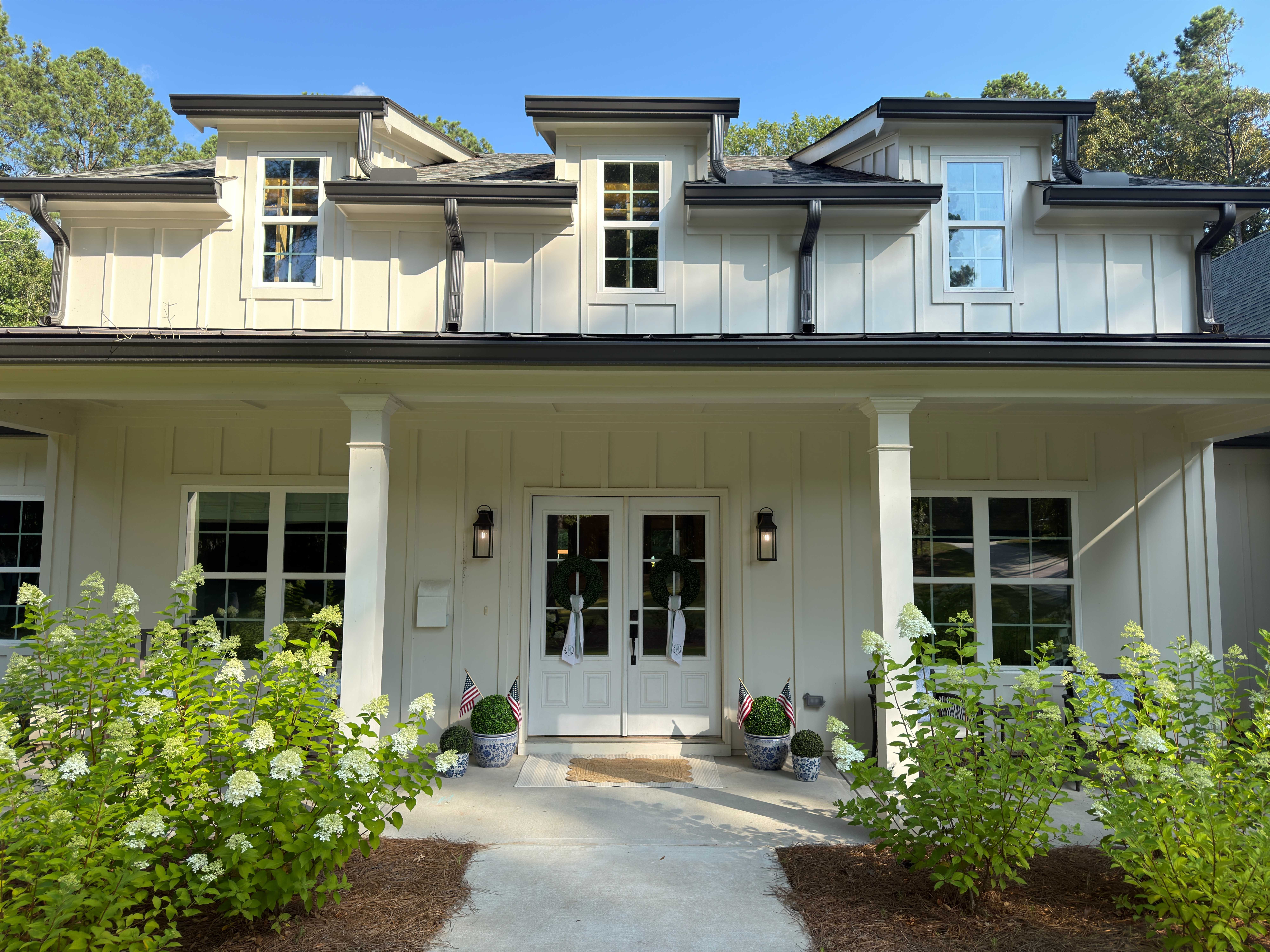 Custom white board-and-batten home with a covered porch, dormers, dark metal roof accents, and hydrangeas along the entry walk