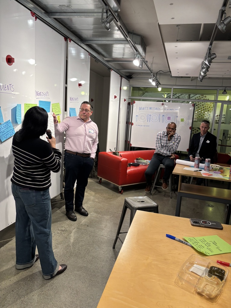 Sunanna presenting R&D Times to group near whiteboard with sticky notes