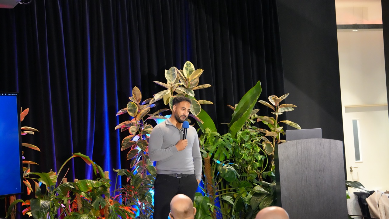 Speaker presenting on the main stage at Cloud Native & Open Source AI Conference with tropical plant backdrop