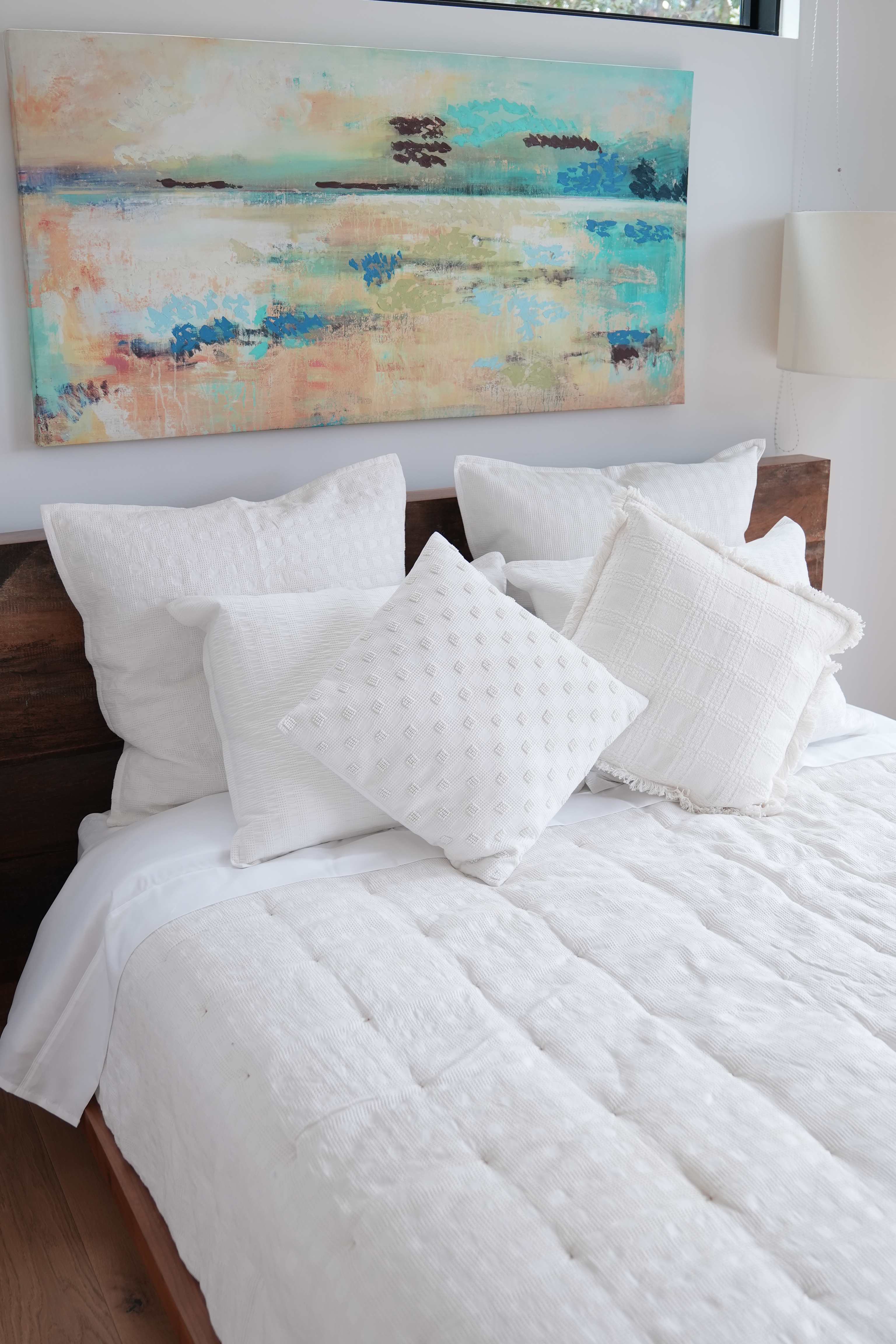 A hotel-style white bed with layered pillows and a neatly made duvet beneath colorful artwork