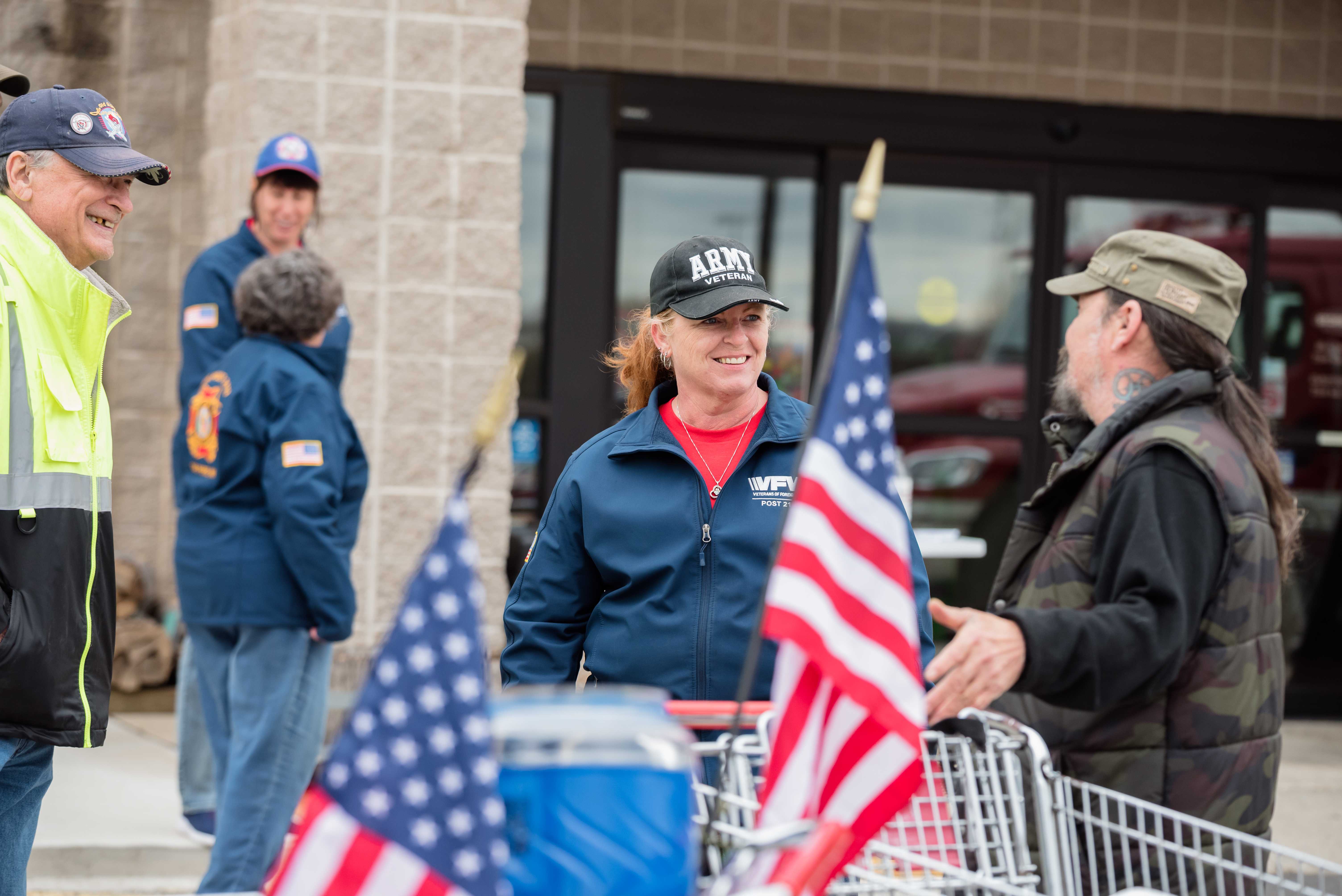 Female Army veteran in VFW jacket smiling at a community fundraising event with American flags