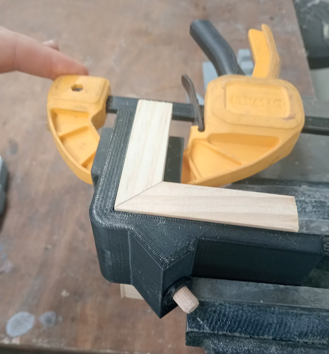 Black 3D printed corner clamping fixture holding two mitre-cut pine pieces at 90 degrees, secured with DeWalt clamps
