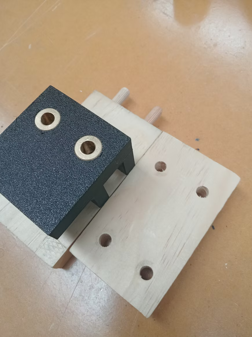 Dark 3D printed dowel drilling jig with brass bushes sitting on a pale timber board with clean dowel holes