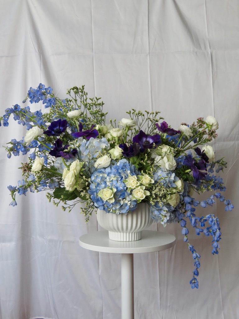 Blue hydrangea, white roses, and purple anemone arrangement in white ribbed bowl