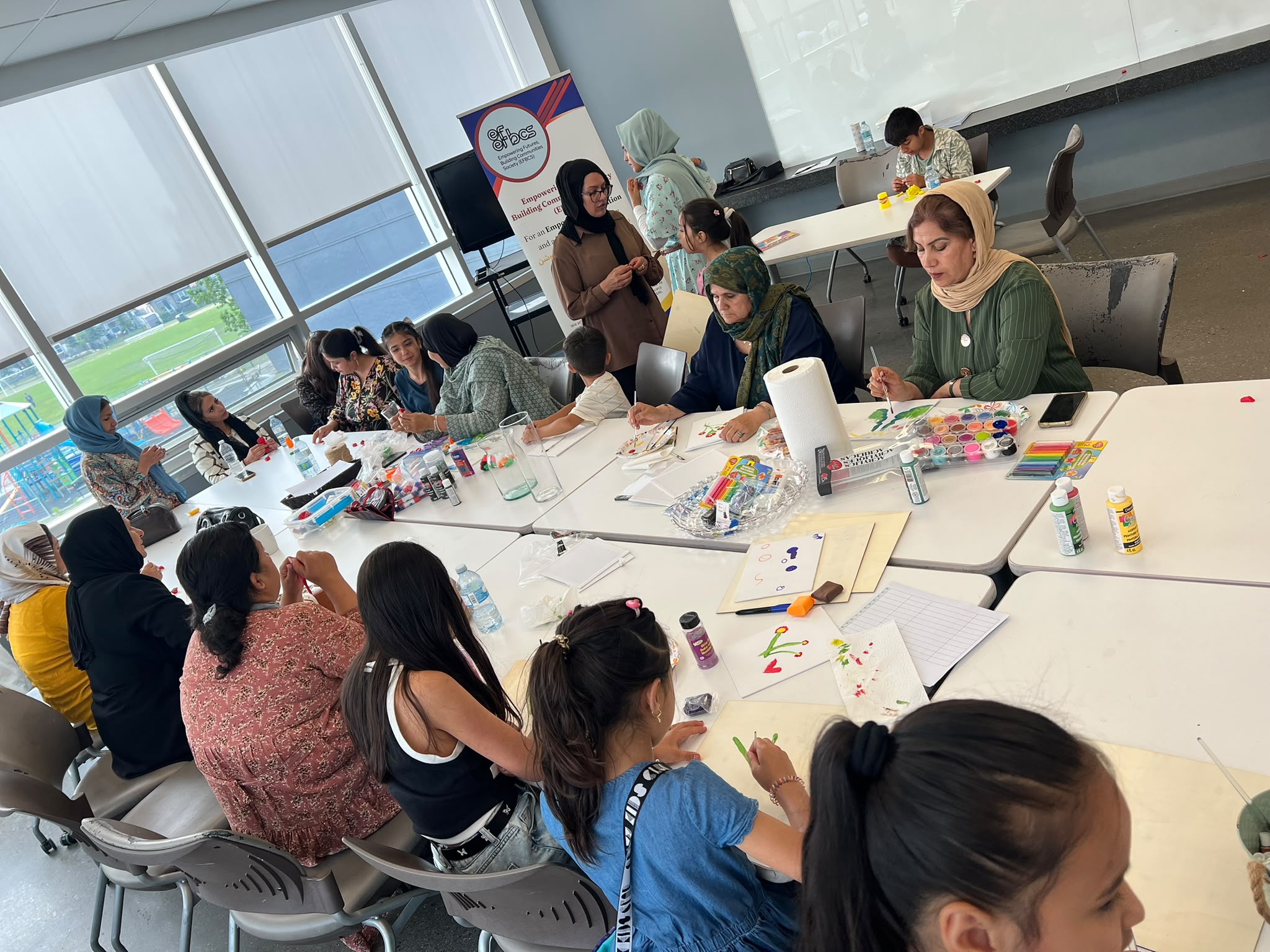 EFBCS arts and crafts workshop — women and children painting together at tables with the EFBCS banner in the background