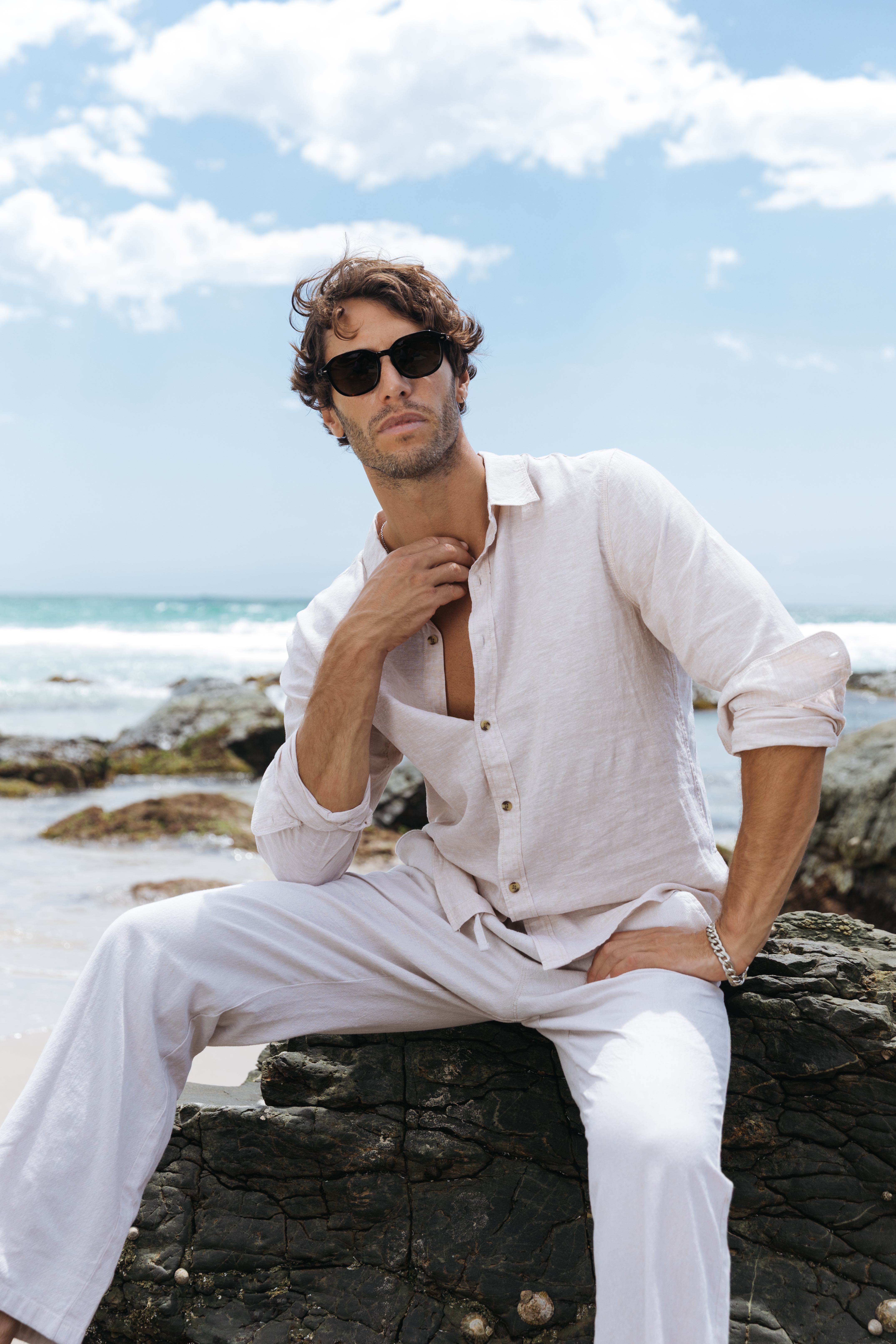 Elias seated on rocks in cream linen shirt white pants