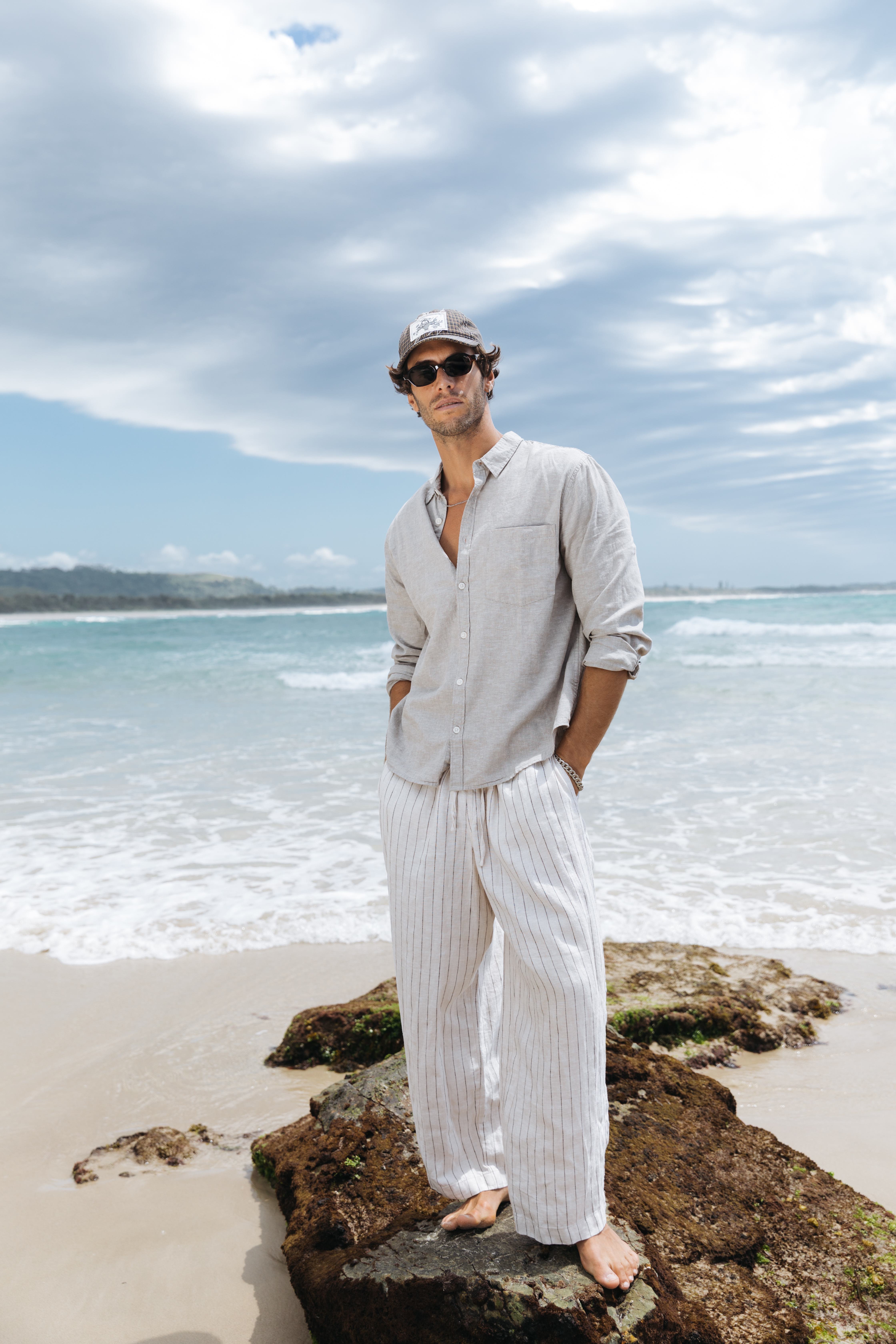 Elias standing barefoot in grey shirt striped linen pants beach