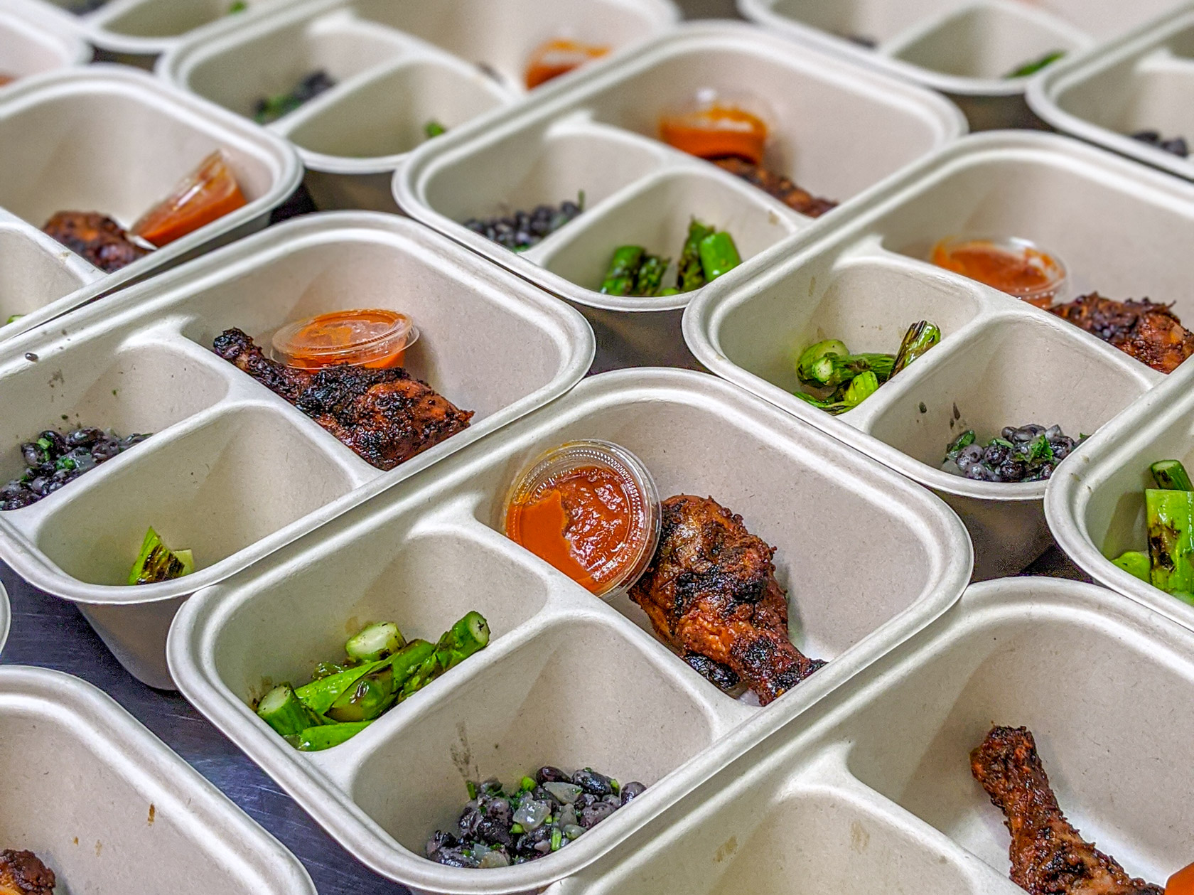 Rows of compostable meal trays filled with grilled chicken, black bean salad, charred asparagus, and cups of sauce