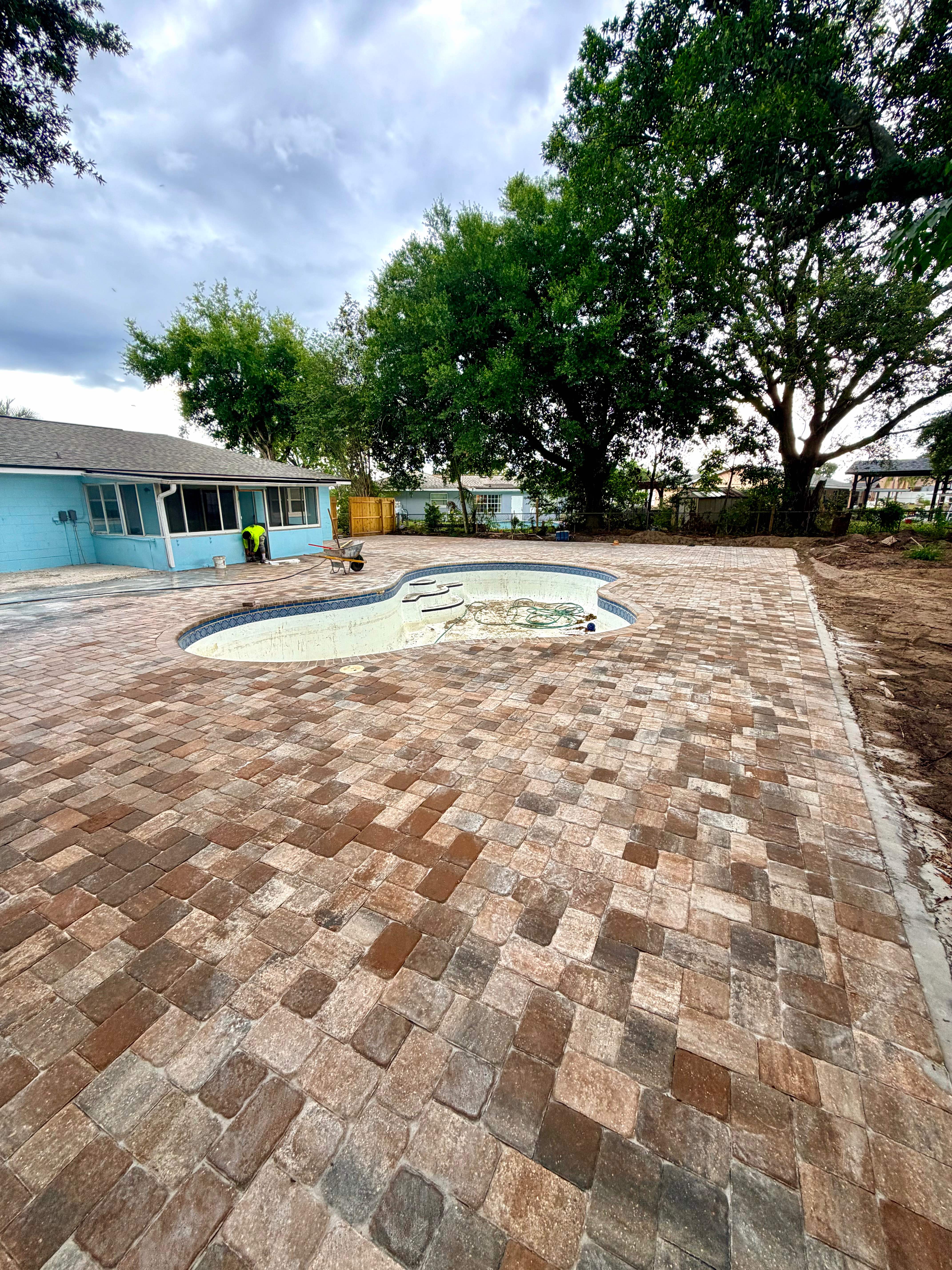Warm terracotta and rust mixed-color cobblestone pavers surrounding an empty kidney-shaped pool with large oak trees overhead