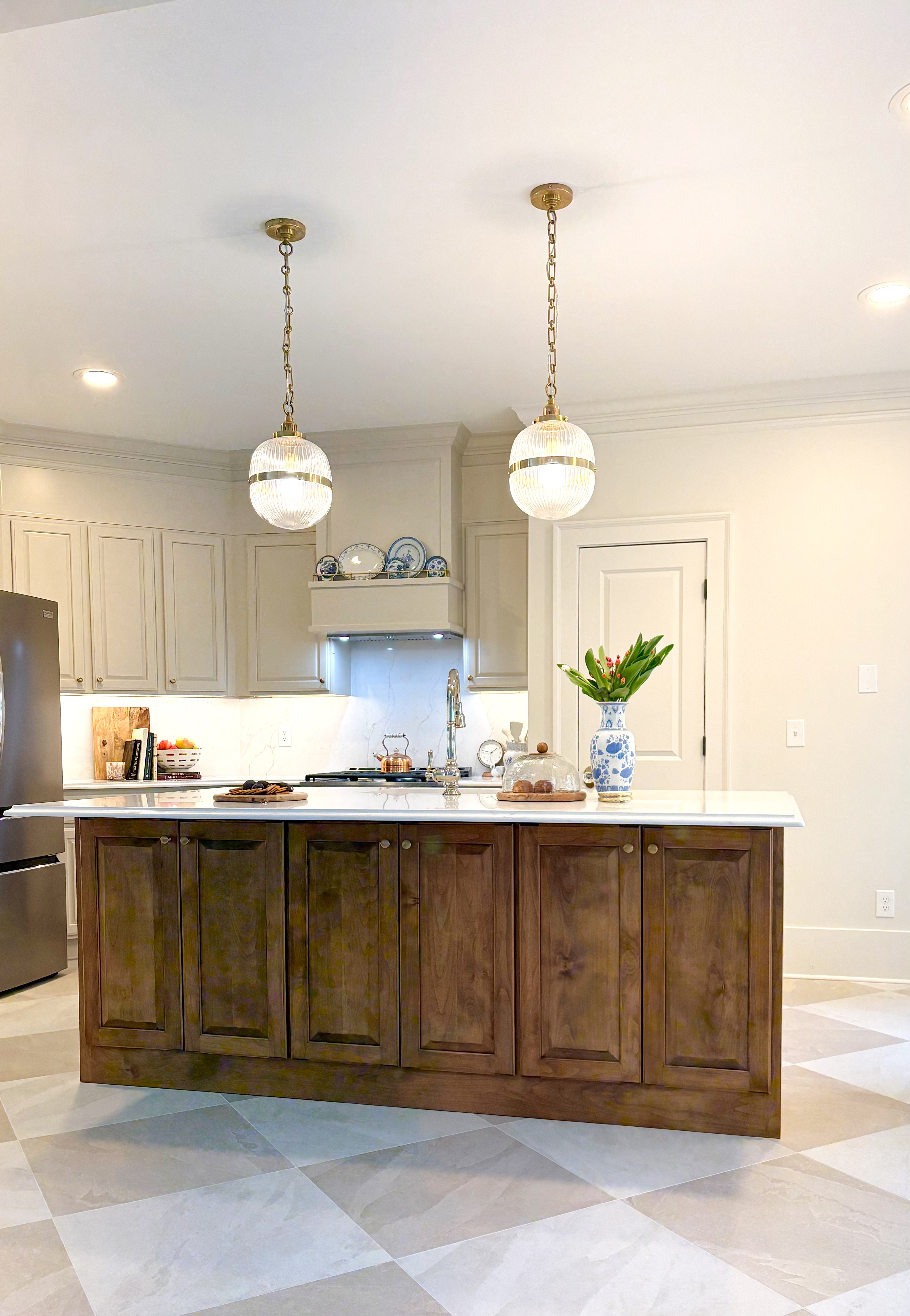 Classic kitchen with a stained-wood island, marble waterfall countertop, twin ribbed-glass globe pendants with brass bands, cream cabinetry, and a gray-and-white checkerboard tile floor