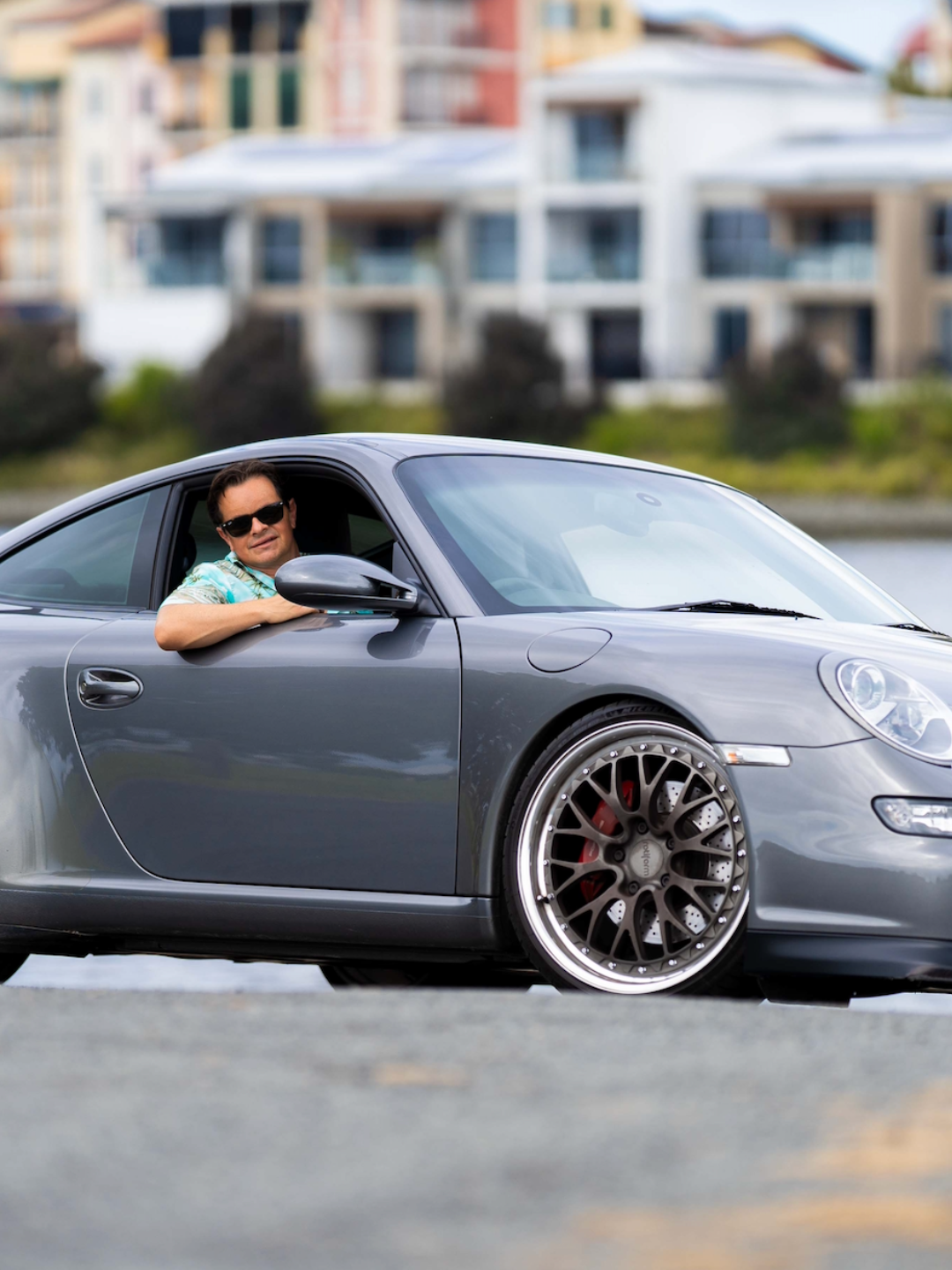 Greg Cassar leaning out of his grey Porsche 911 with aftermarket wheels, wearing sunglasses, with waterfront buildings in the background.