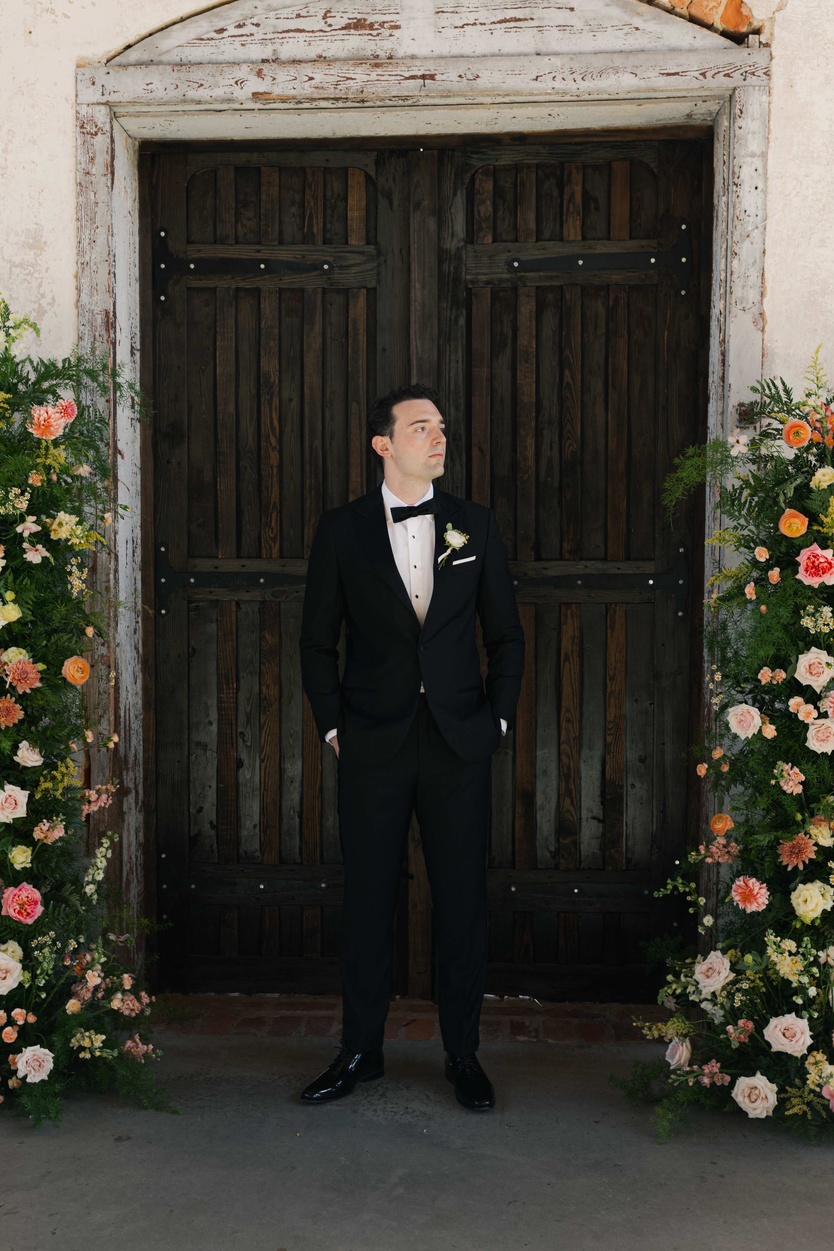 Groom in a black tuxedo framed by tall wooden doors and floral installations