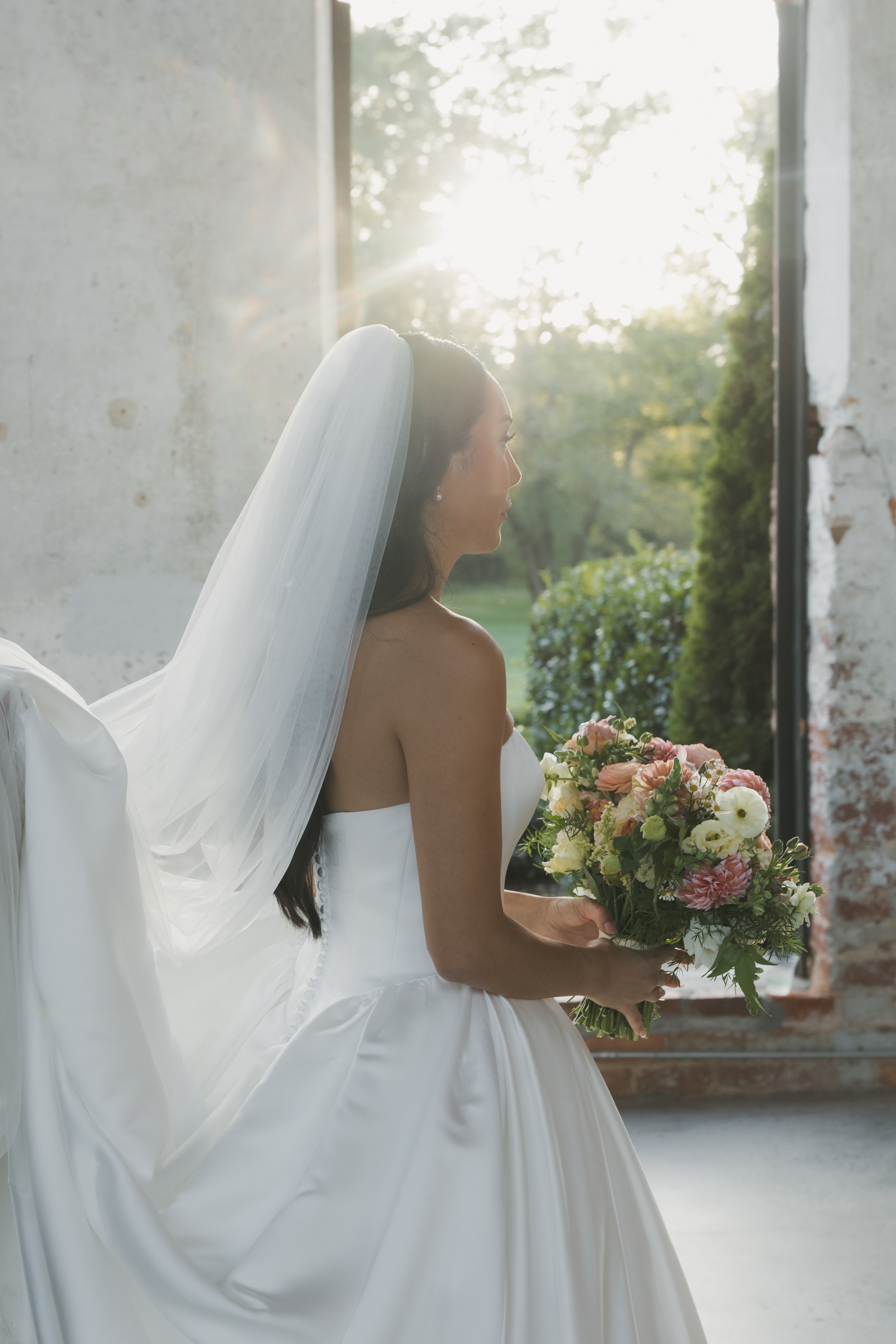 Bride with a long veil backlit by golden light, holding a pastel garden bouquet