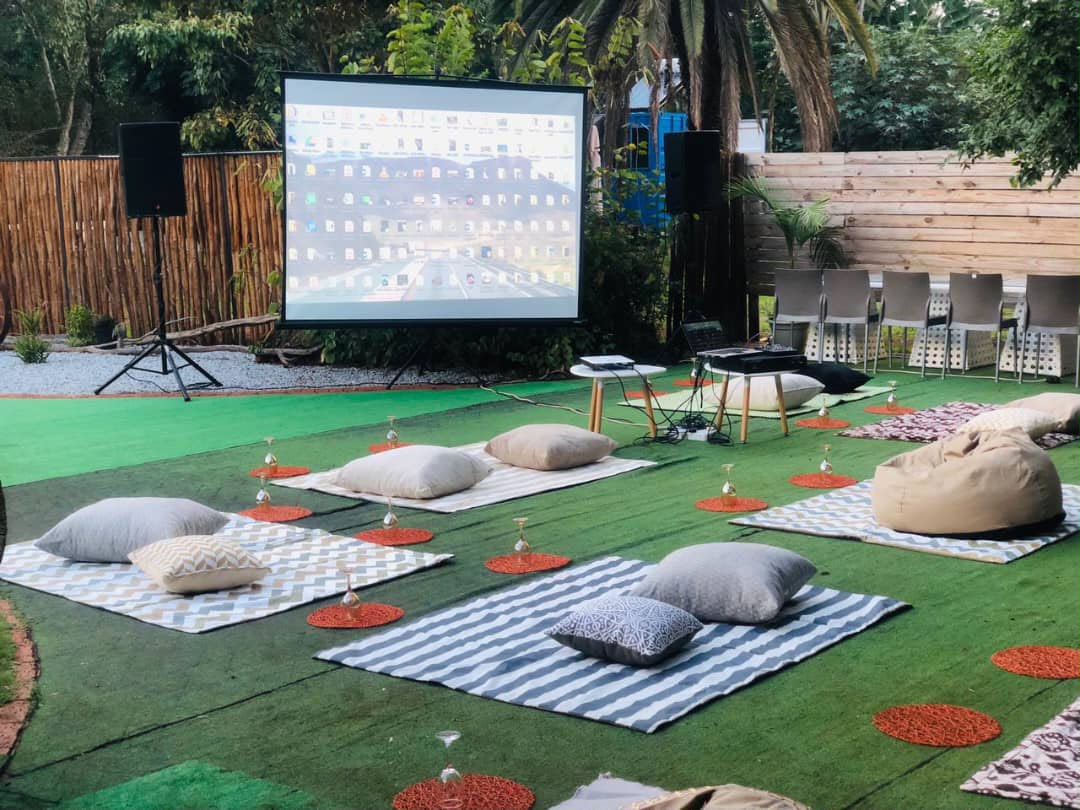 Outdoor garden event setup with projection screen, beige cushions, DJ equipment, professional staging