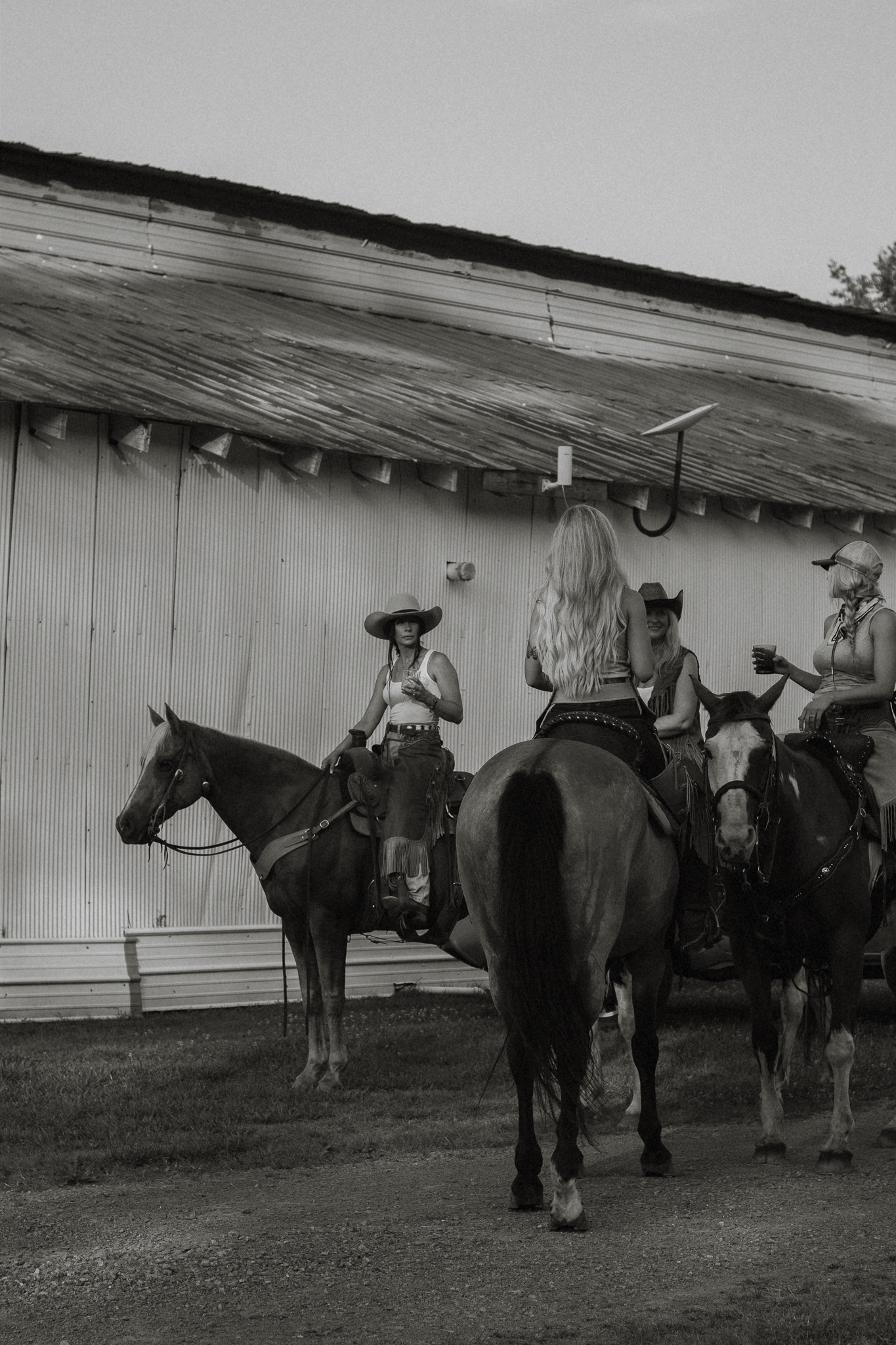 Cowgirls on horseback gathered by barn in black and white