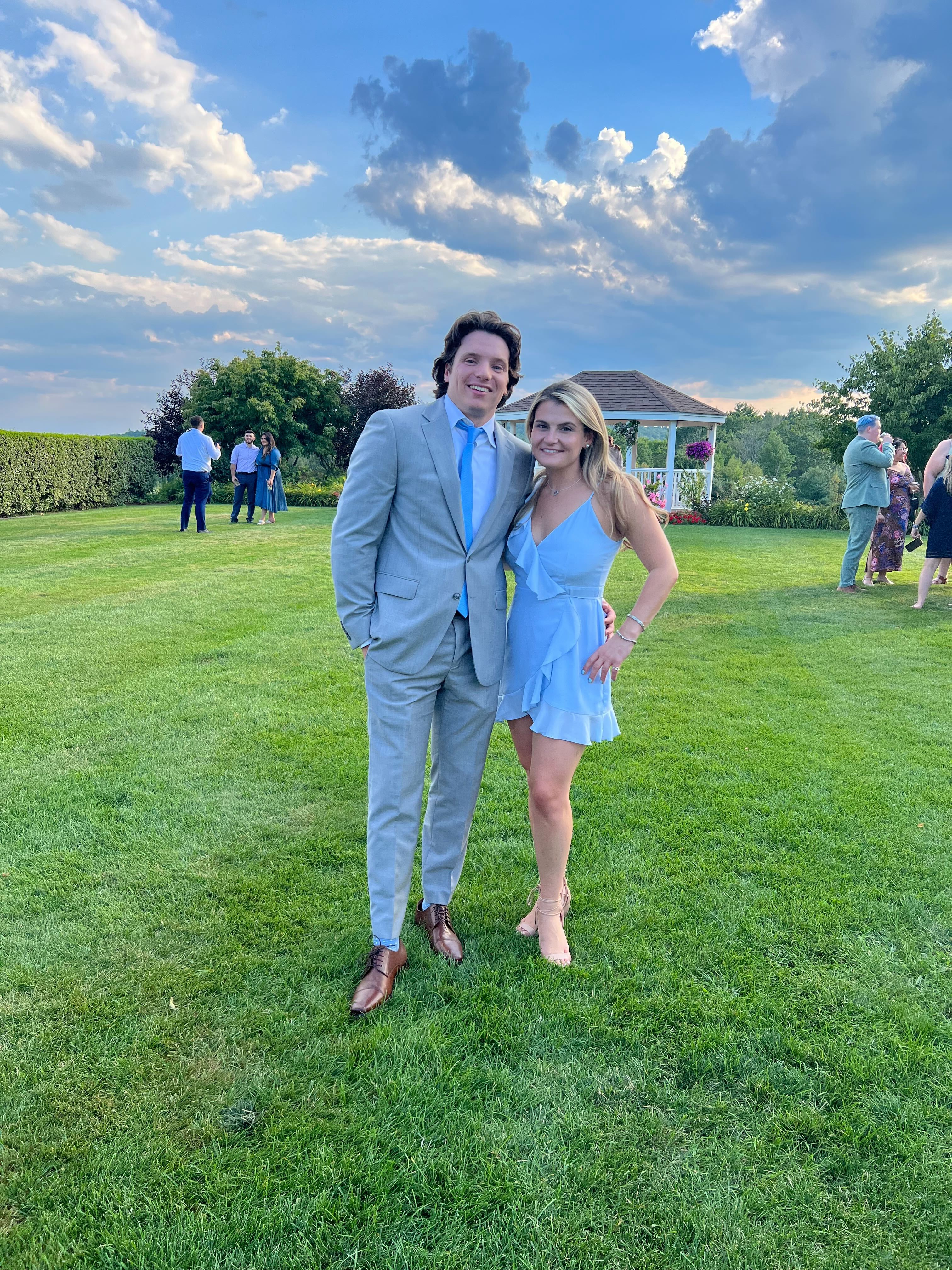 Ben and Brooke at an outdoor event, Ben in a light grey suit and Brooke in a light blue dress, with a garden and gazebo in the background