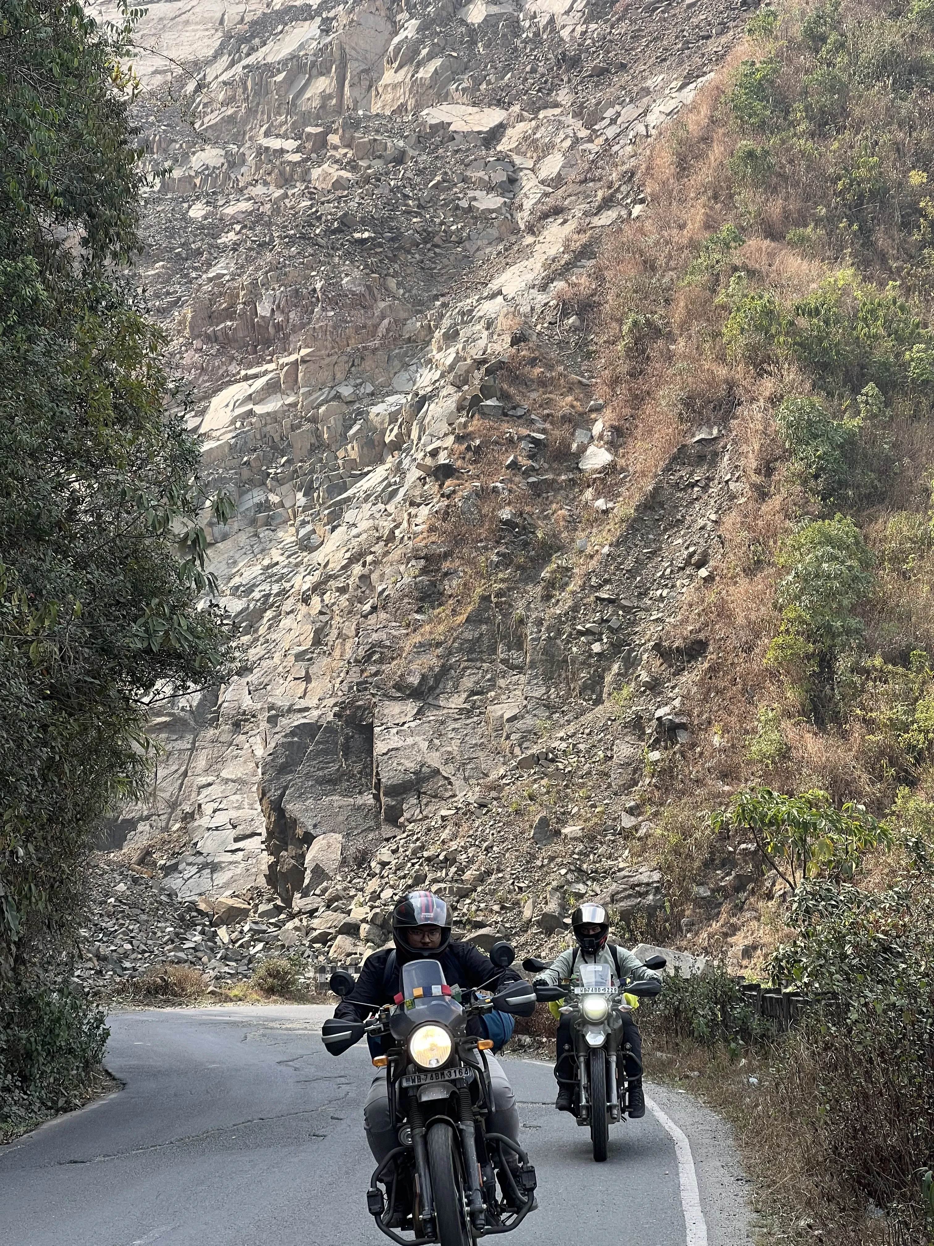 Two adventure motorcyclists riding Royal Enfield Himalayan 411 and Hero XPulse 200 2V along a mountain road with impressive Himalayan rock formations and cliff faces, demonstrating group touring capabilities. Premium bike rentals in North Bengal for group riding adventures.