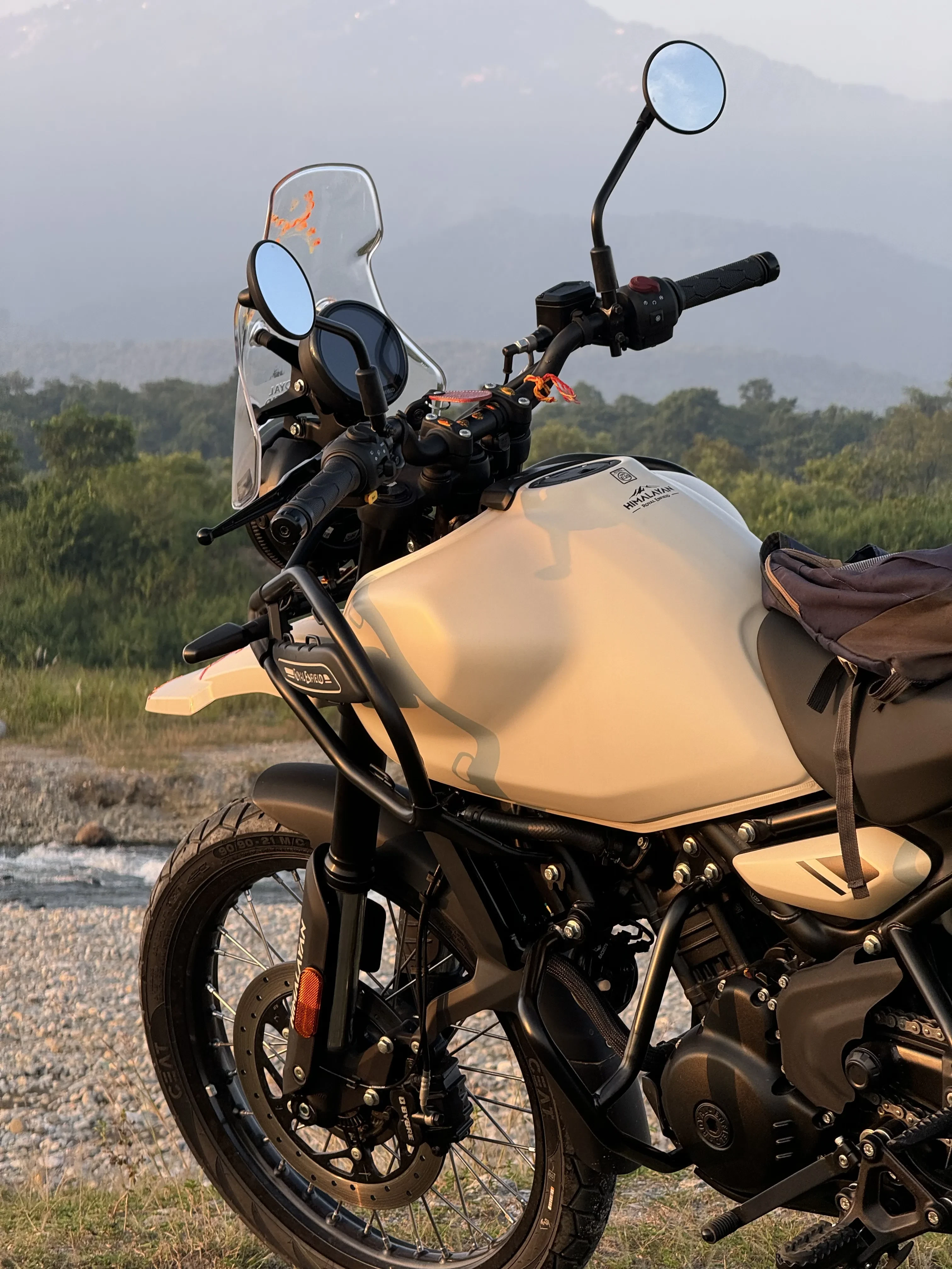 Royal Enfield Himalayan 450 Kaza Brown motorcycle (in sand colour) parked by a riverbed with majestic Himalayan mountain ranges in background during golden hour, highlighting the perfect motorcycle for mountain exploration. Affordable bike rentals in Sikkim for Himalayan exploration.