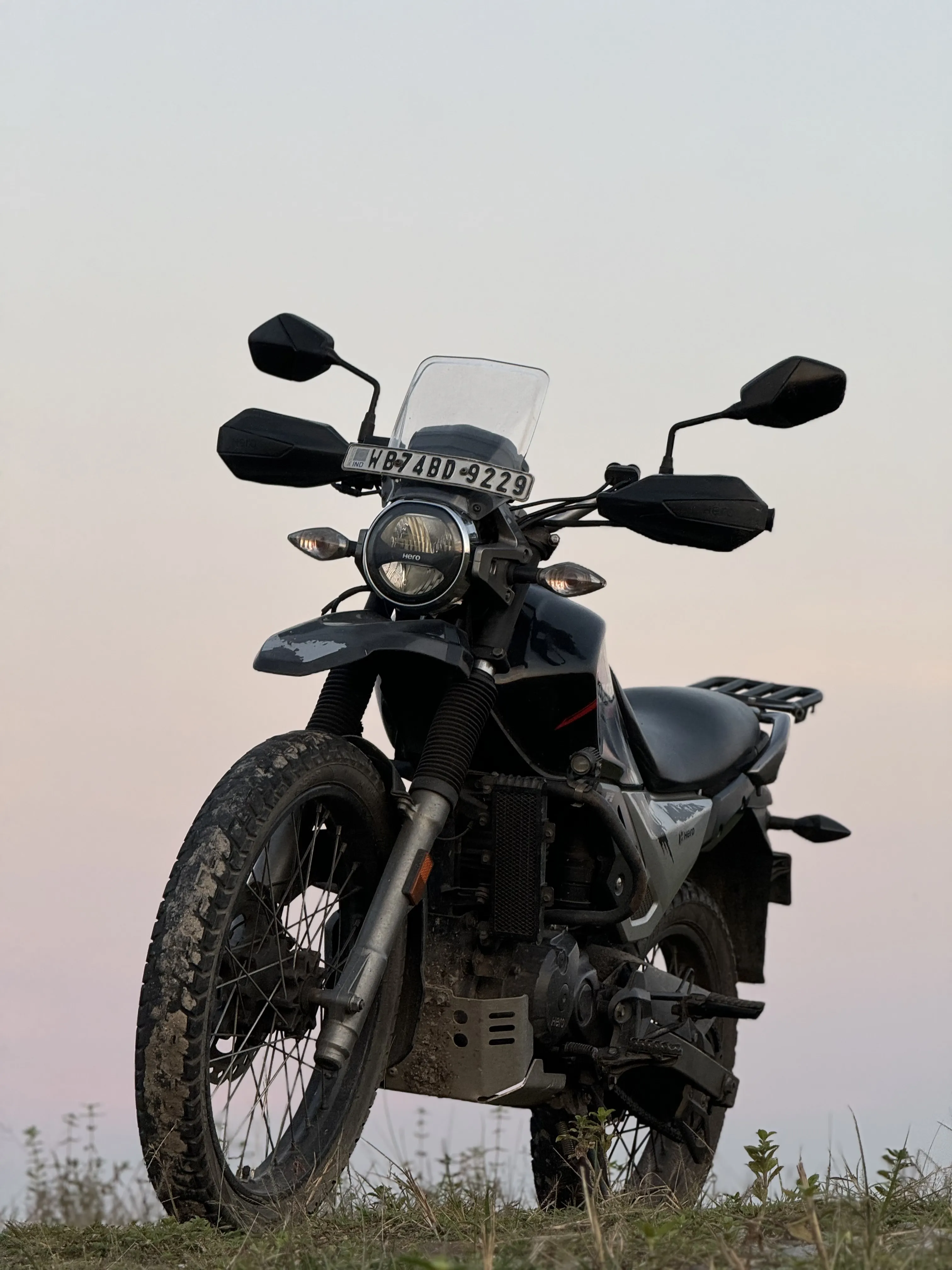 Hero XPulse 200 2V adventure motorcycle front view with rugged off-road tires and protective hand guards positioned on grassy terrain at dusk, showcasing its robust build designed for Himalayan terrain exploration. Premium motorcycle rentals in Darjeeling for off-road adventures.