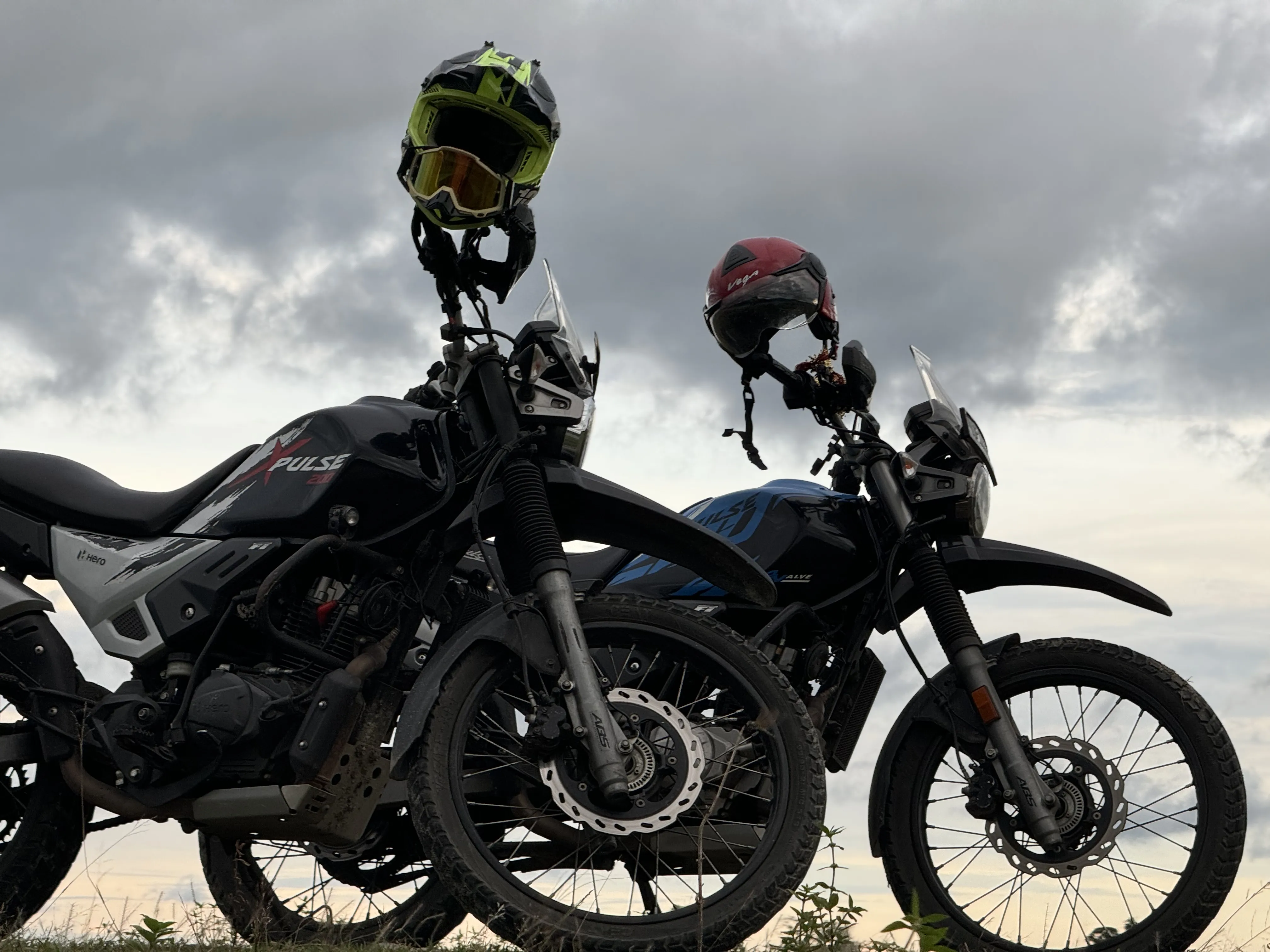 Two Hero XPulse adventure motorcycles parked side by side with helmets mounted on handlebars against dramatic cloudy Himalayan sky at sunset, ready for group touring adventures. Premium motorcycle rentals in Siliguri for group rides.