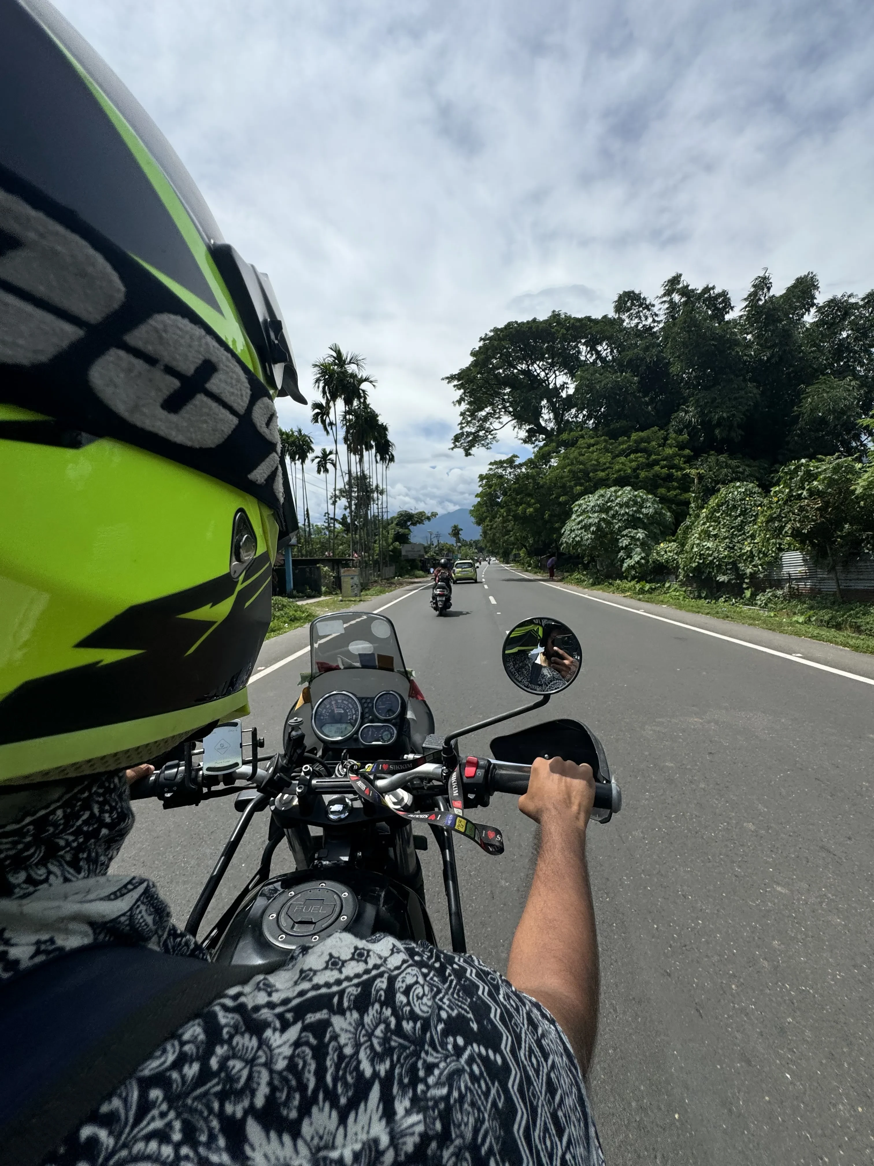 First-person rider perspective from a Royal Enfield motorcycle on a smooth Himalayan highway with lush tropical vegetation and palm trees lining the route, showcasing the rider experience with neon safety helmet. Affordable motorcycle rentals in Bagdogra Airport for highway cruising.