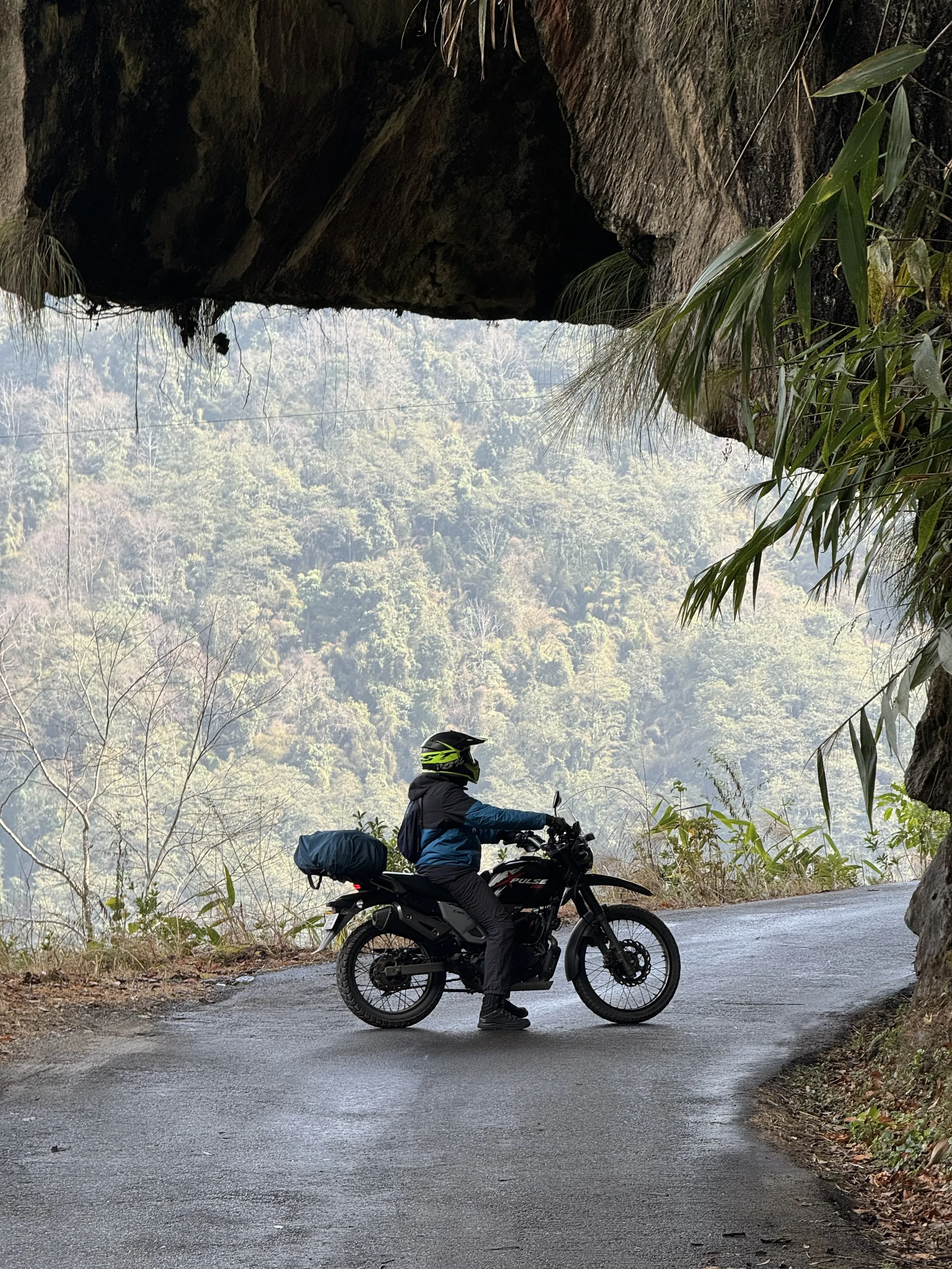 Adventure motorcyclist stopped under a dramatic rock overhang on a mountain road with panoramic valley views, showcasing the hidden gems accessible by motorcycle in the Himalayas. Premium motorcycle rentals in Bagdogra Airport for scenic viewpoints.