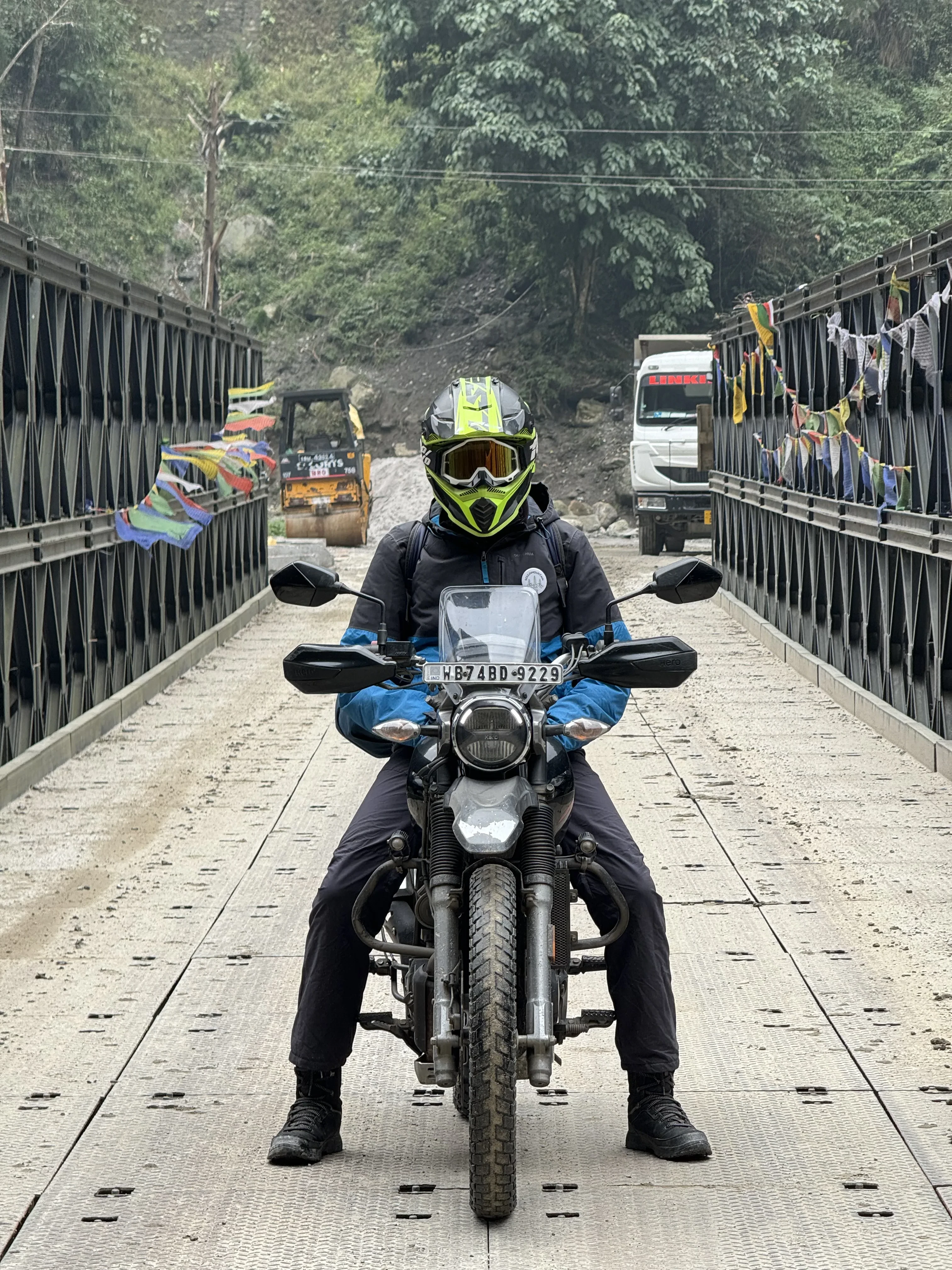 Adventure motorcyclist in full safety gear riding a Hero XPulse 200 2V on a metal bridge crossing in the remote Himalayas, with colorful prayer flags and construction equipment visible in the mountainous background. Affordable motorcycle rentals in Siliguri for remote Himalayan exploration.