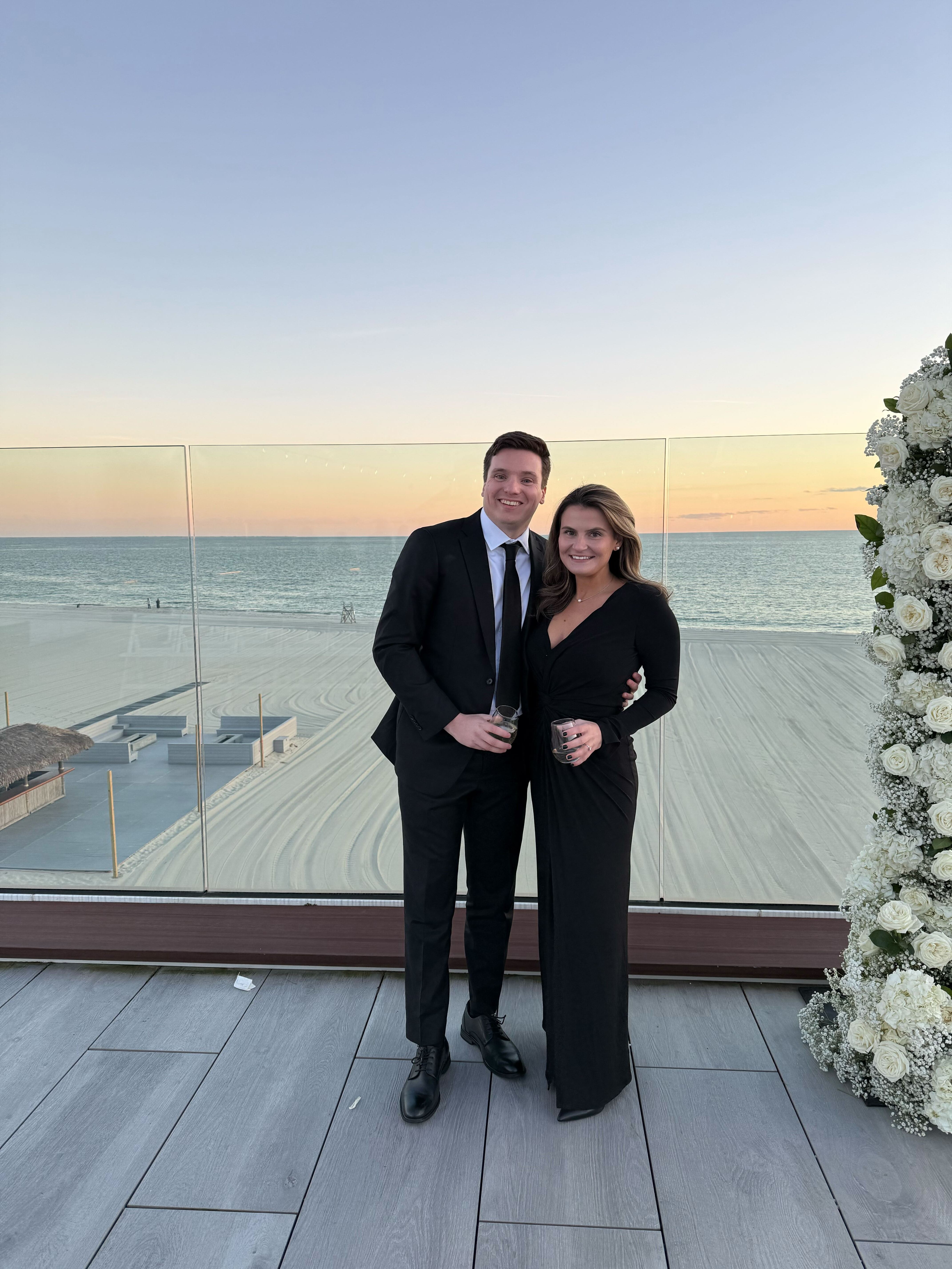 Ben and Brooke in formal attire standing on a balcony overlooking the ocean at sunset