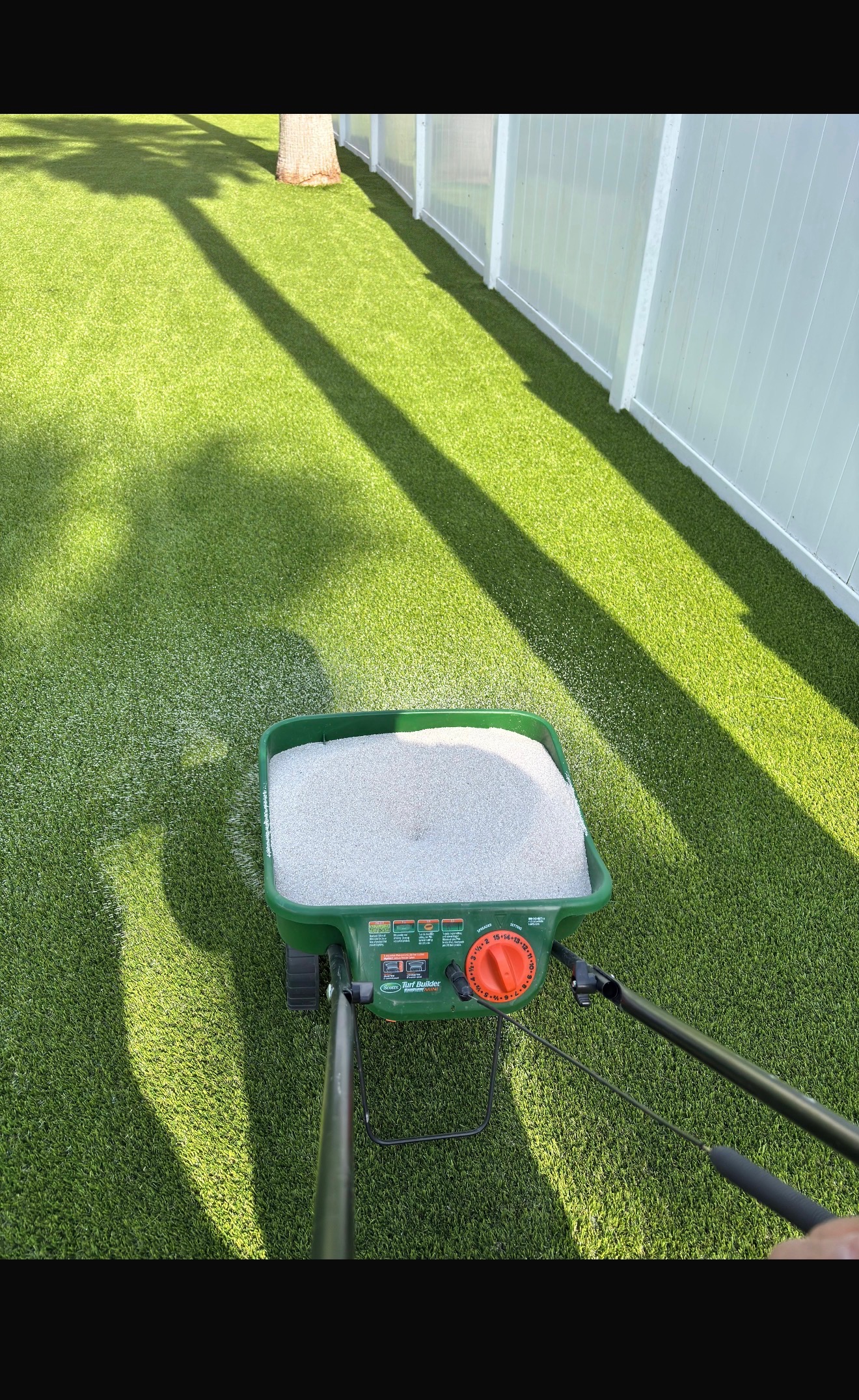Silica infill sand broadcast-spread across freshly installed artificial grass beside a white vinyl fence