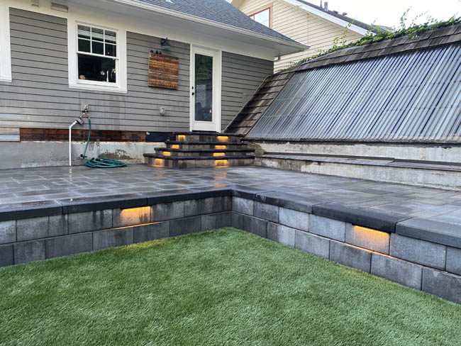 Two-tier charcoal paver patio with step lighting beside synthetic turf
