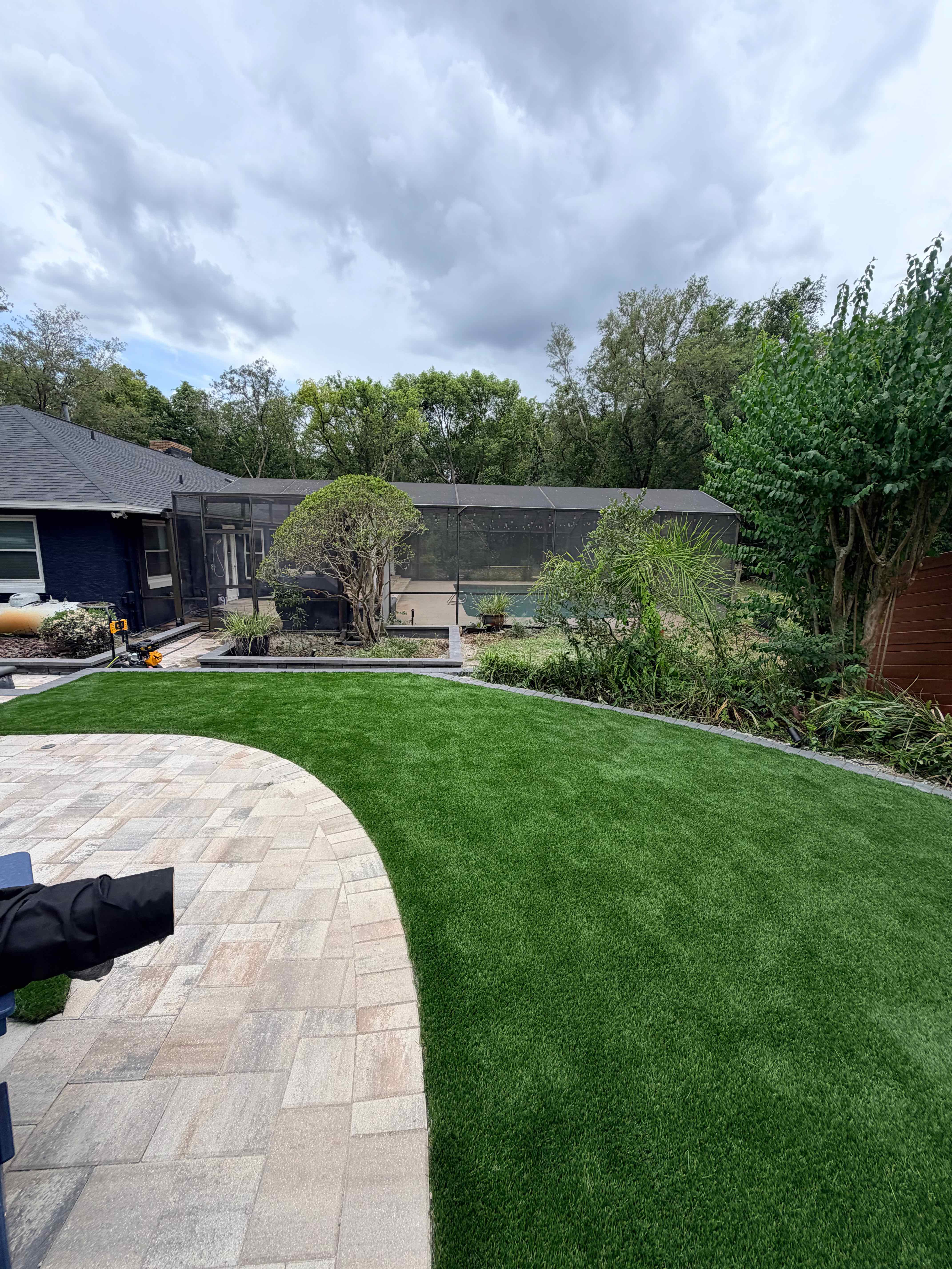 Curved artificial grass lawn with large travertine paver patio and lush garden beds beside screened enclosure