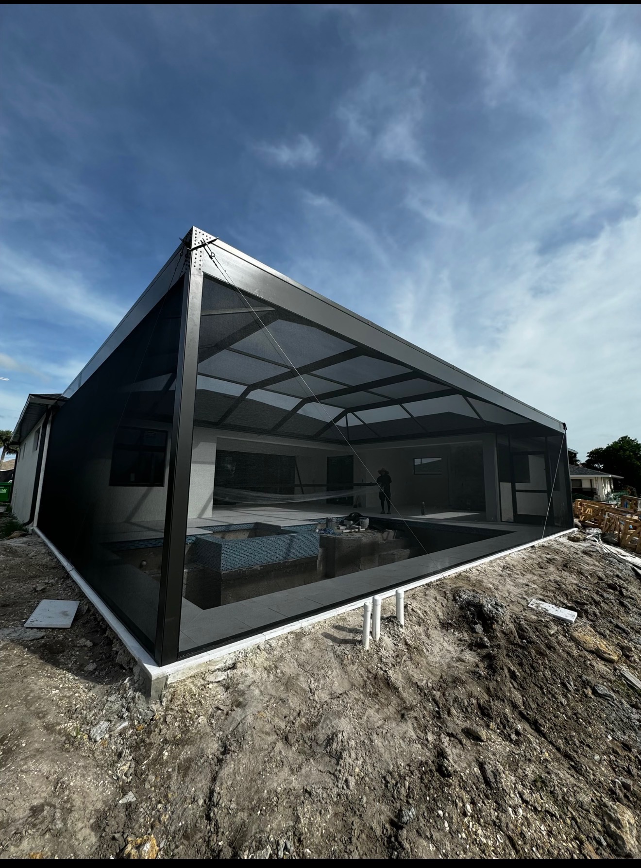 Large angular modern pool enclosure under construction with dark charcoal aluminum frame and dramatic triangular roofline against blue sky