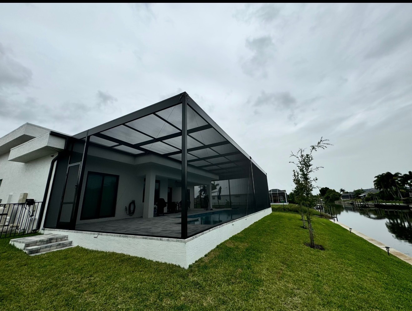 Contemporary flat-roof screen enclosure on a modern white stucco waterfront home with dark aluminum frame and canal view