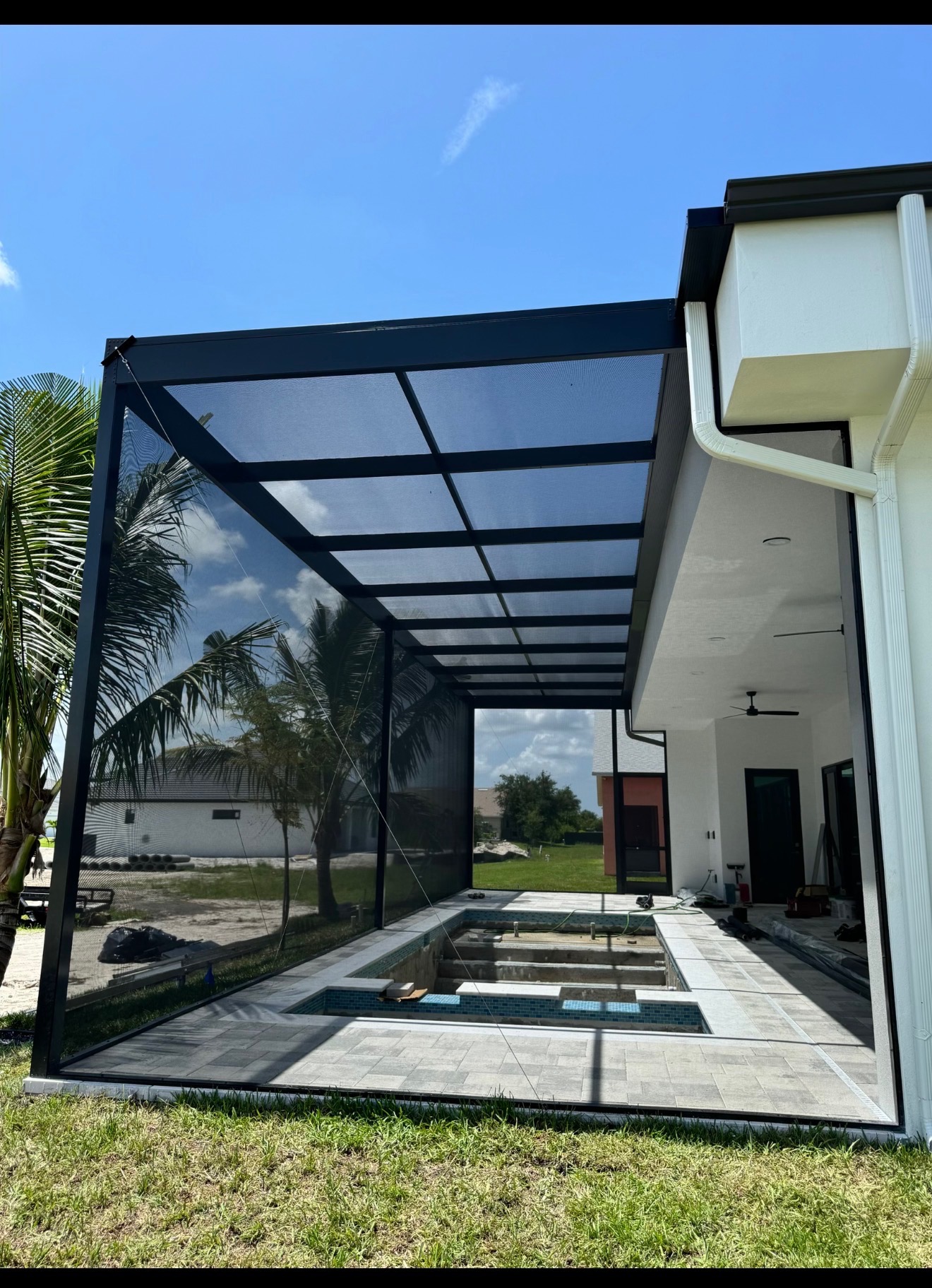Dark aluminum flat-grid screen enclosure attached to a white modern home with pool and spa under construction and travertine deck