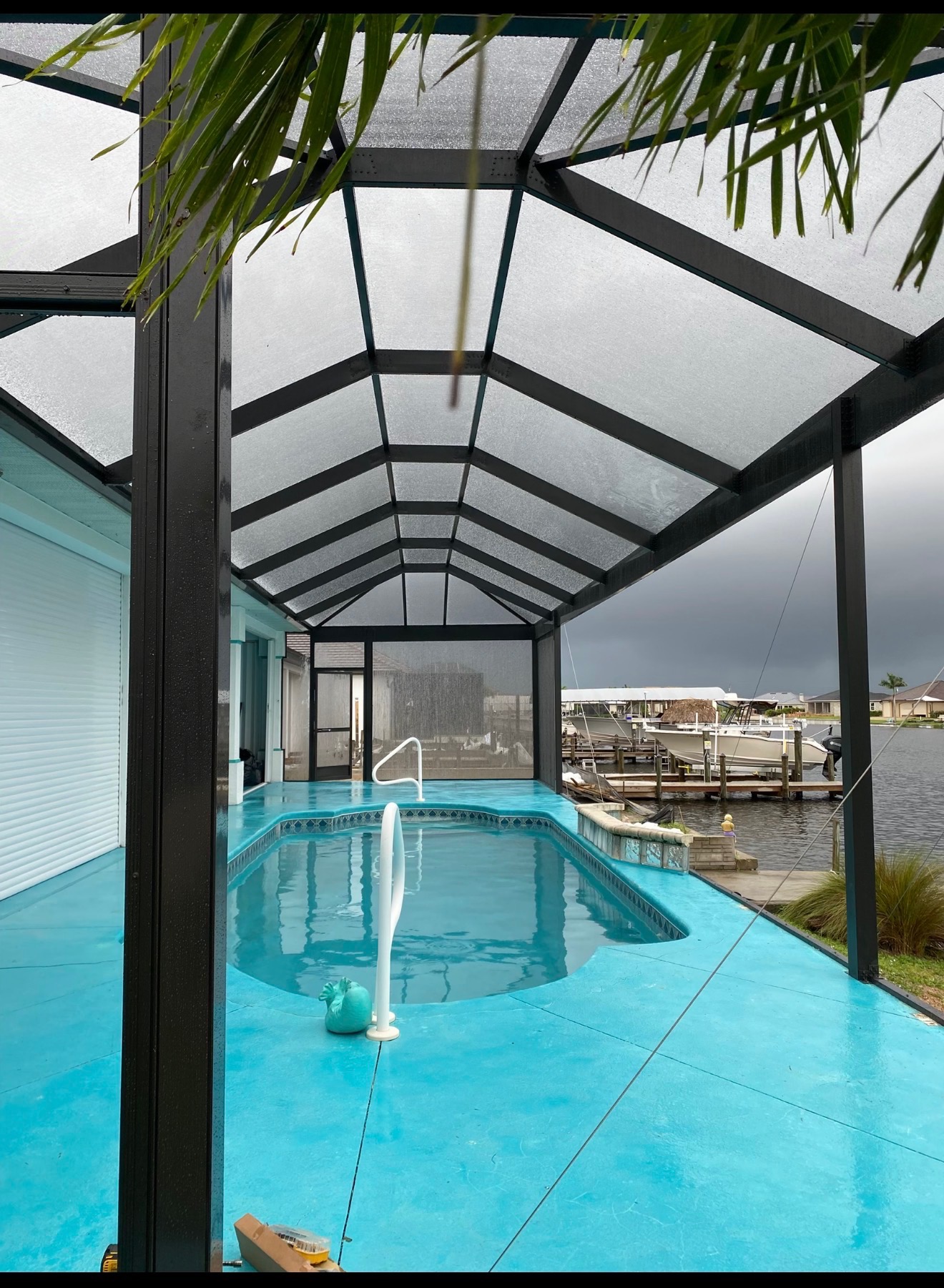 Interior view of a gabled pool enclosure with dark aluminum frame, turquoise pool deck and marina beyond under a stormy sky