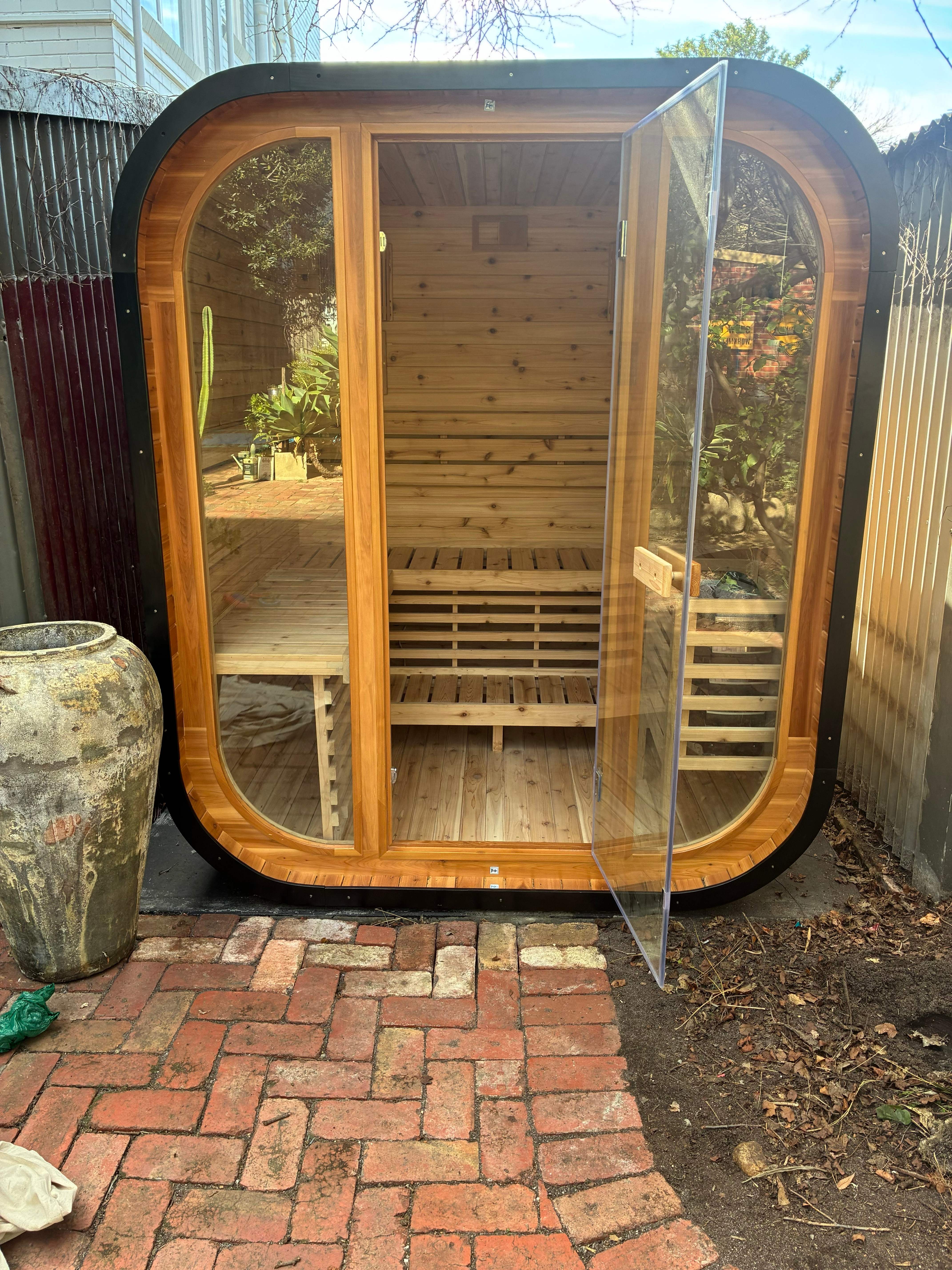 Finished cedar sauna with its glass door open, installed on brick paving in a Melbourne backyard
