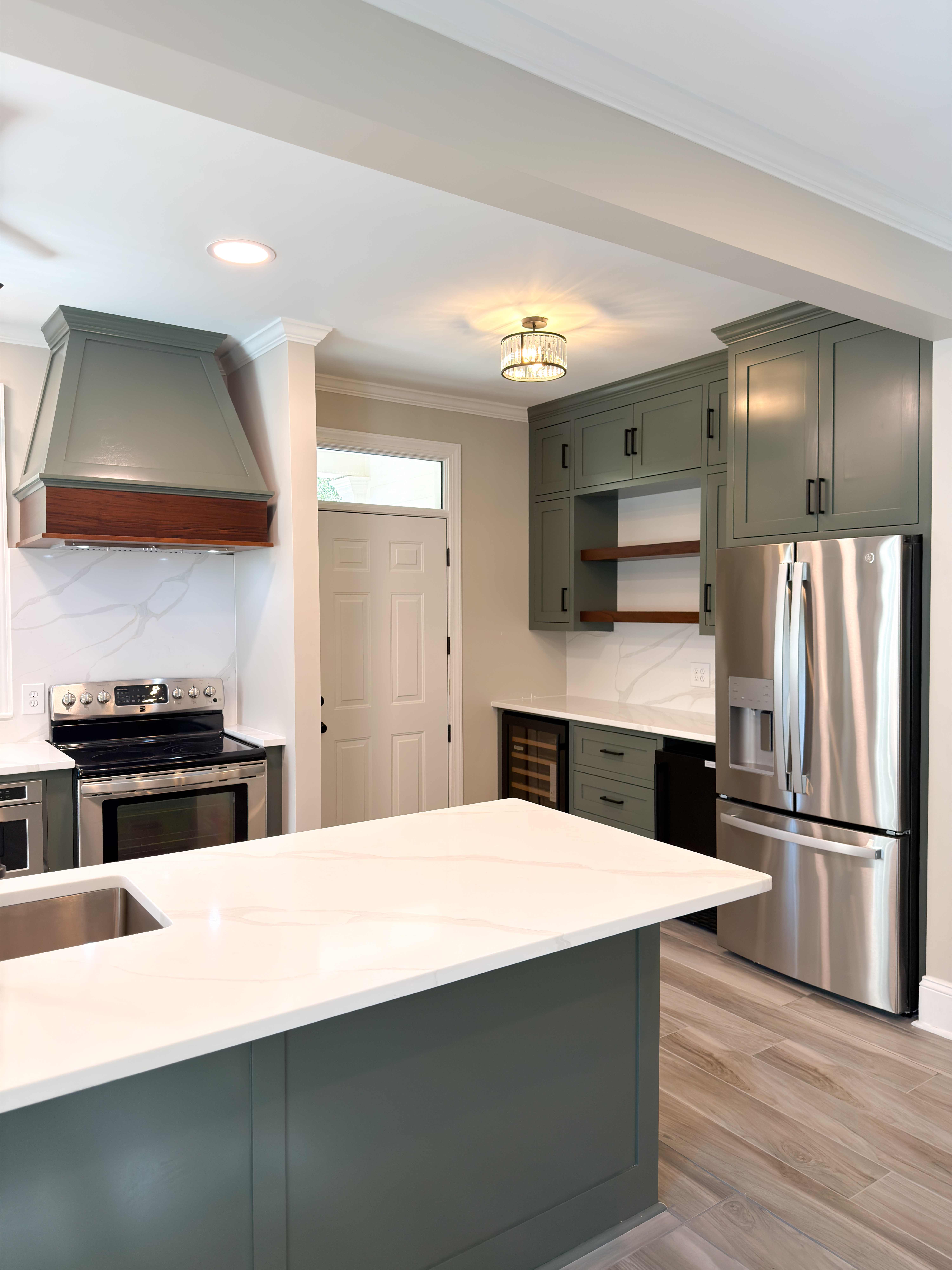 Sage-green kitchen with a marble waterfall island, a green range hood accented with a walnut band, open wood shelving, and a stainless refrigerator over wood-plank floors