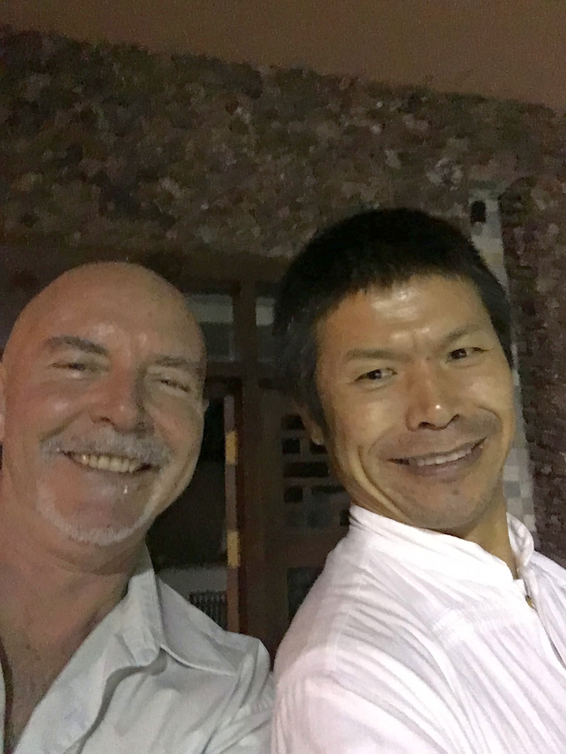 Joyful close-up portrait of Kadama smiling broadly with his Chinese qigong friend, both in white shirts, warm interior lighting