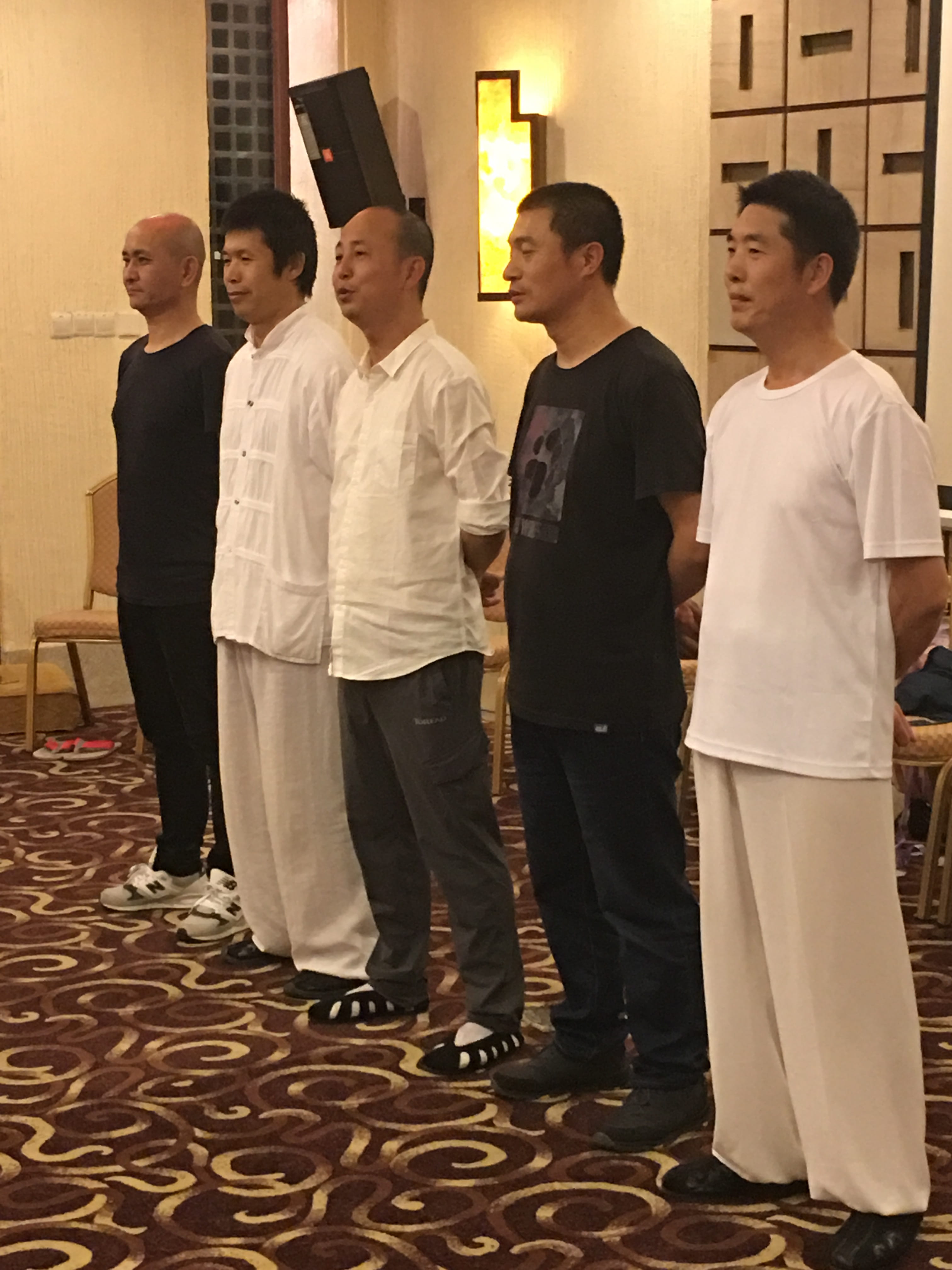 Five Chinese qigong practitioners and Kadama's classmates standing in formal line-up at a ceremonial venue with warm wood walls and patterned carpet
