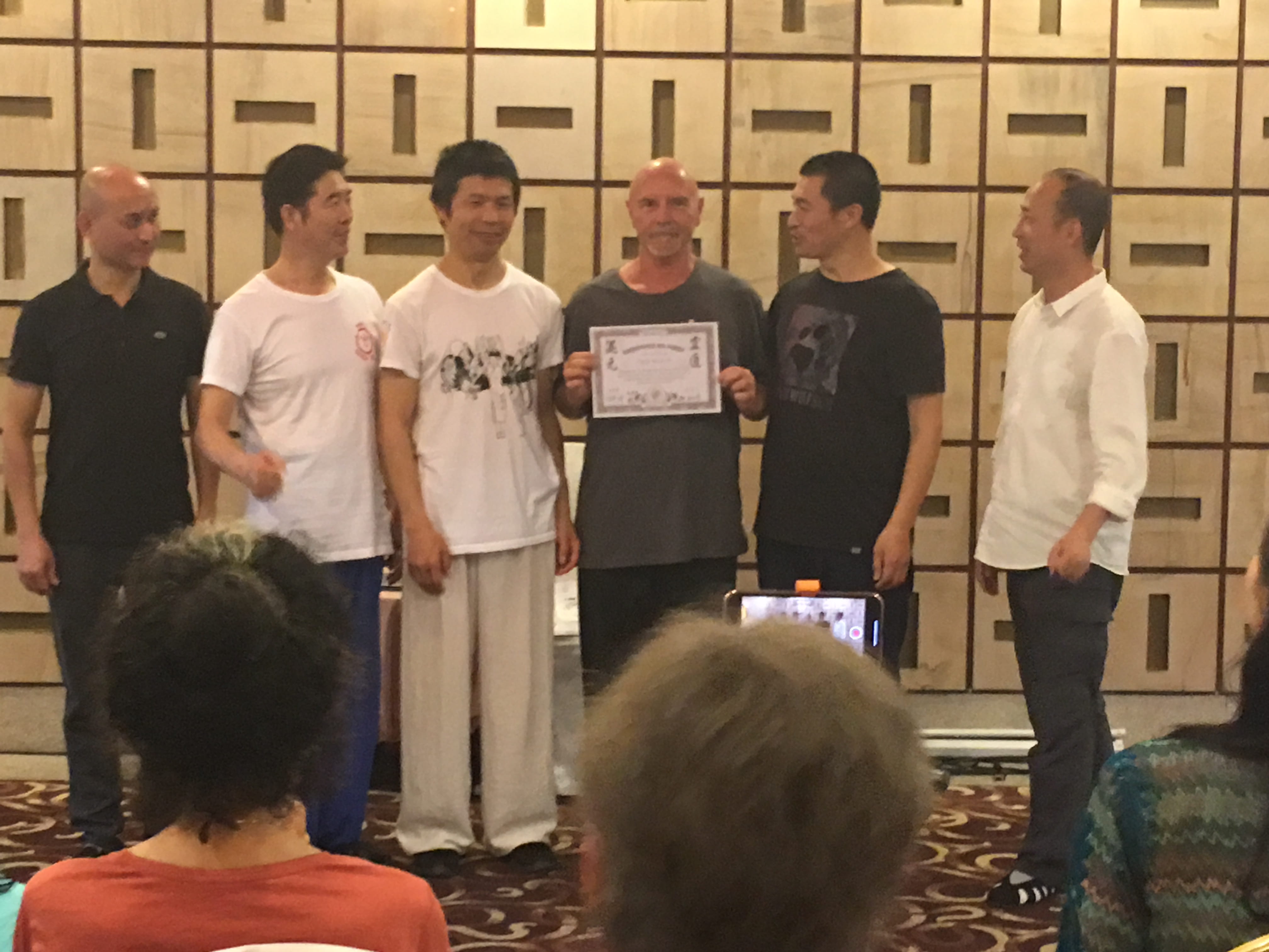 Kadama receiving a Chinese qigong lineage certificate from masters in a ceremonial line-up, six men in front of a warm wood-panelled wall