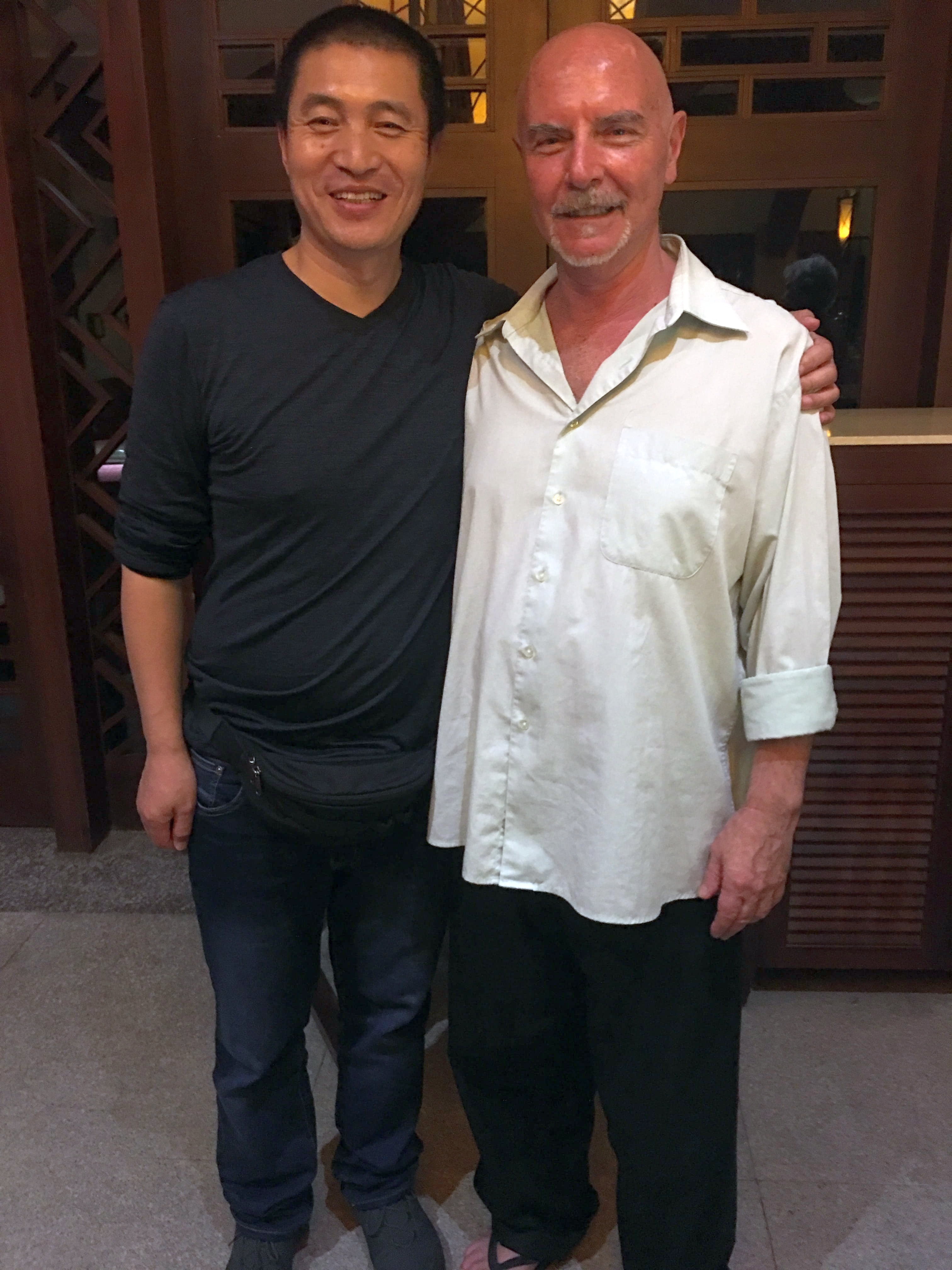Kadama in cream shirt arm-in-arm with his Chinese qigong teacher, standing together in front of warm wooden door panels