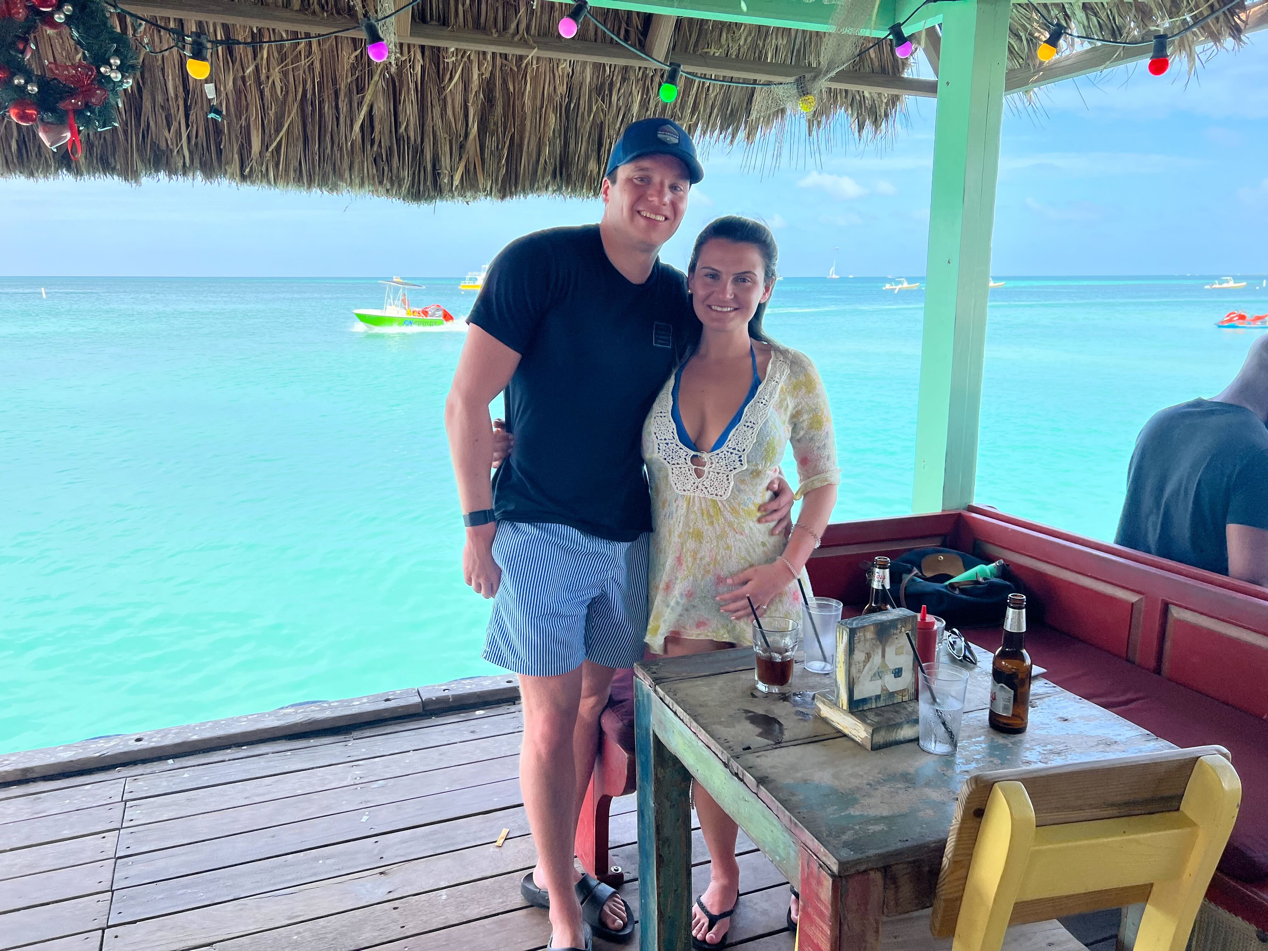 Ben and Brooke at a tropical beach bar with thatched roof and turquoise waters in the background