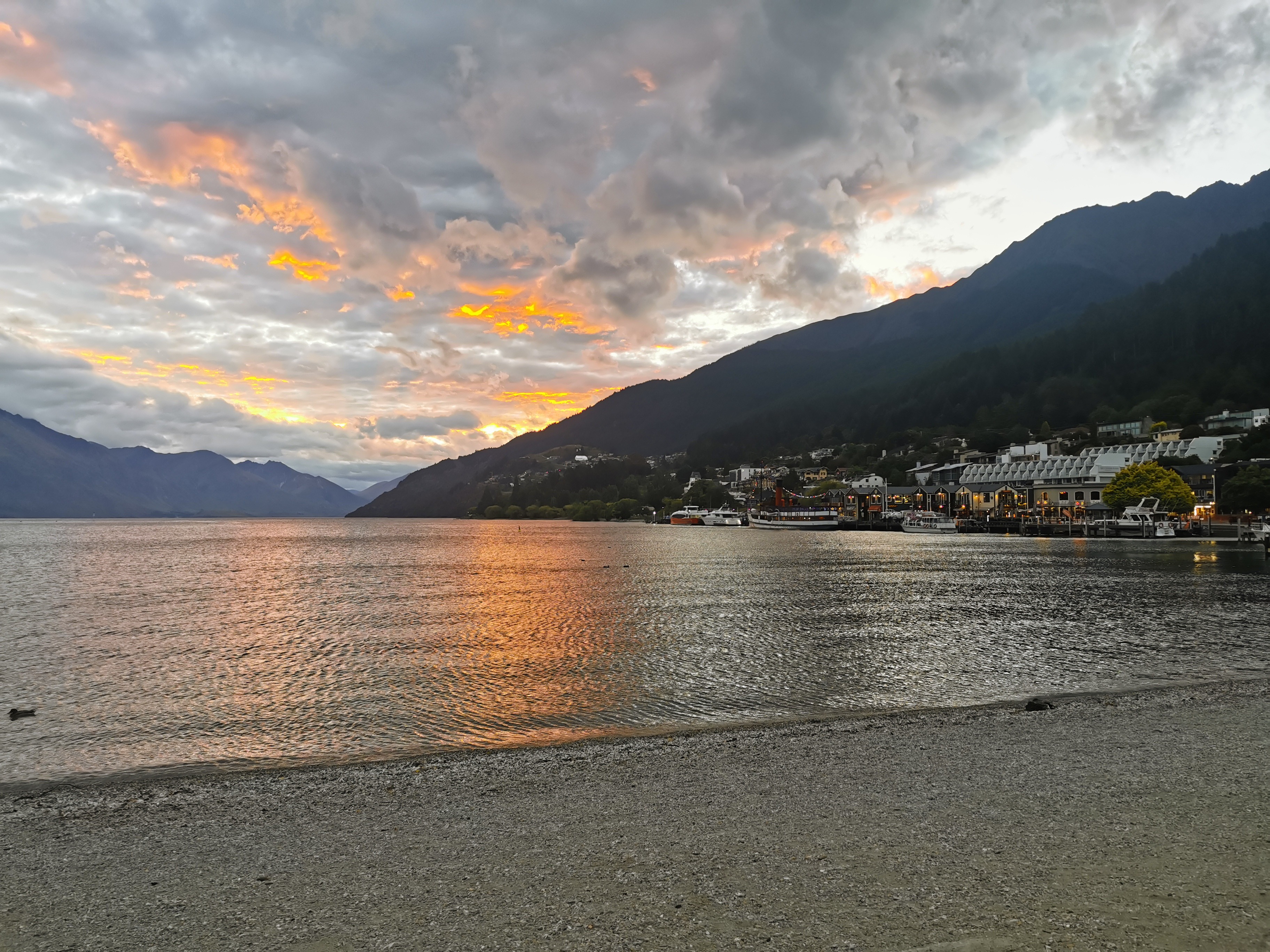Queenstown waterfront at sunset — the Remarkables silhouetted against an orange sky, boats moored along the lakefront
