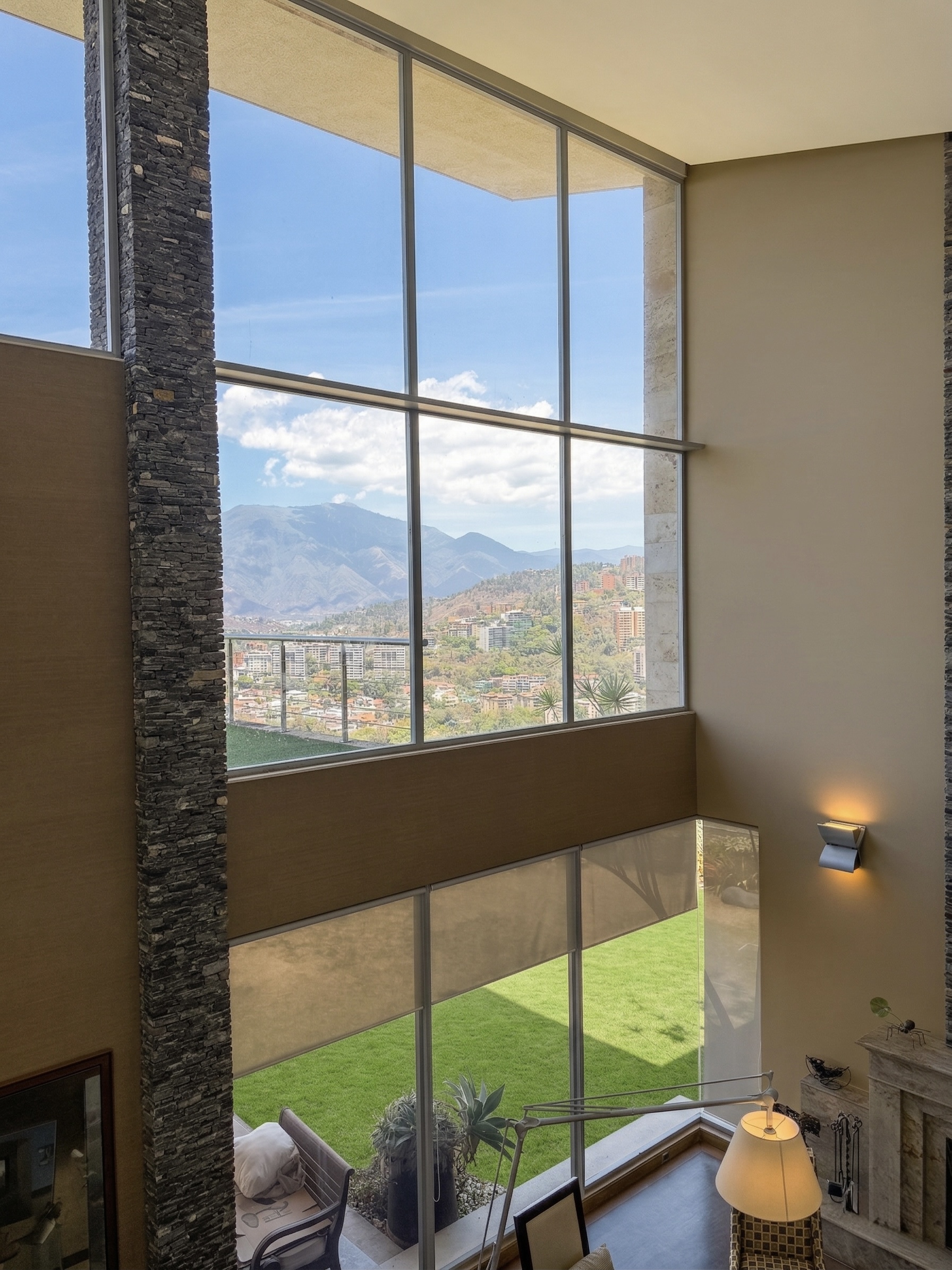 Dramatic double-height living space with floor-to-ceiling windows and panoramic Avila mountain views, stone column detail, La Trinidad estate Caracas