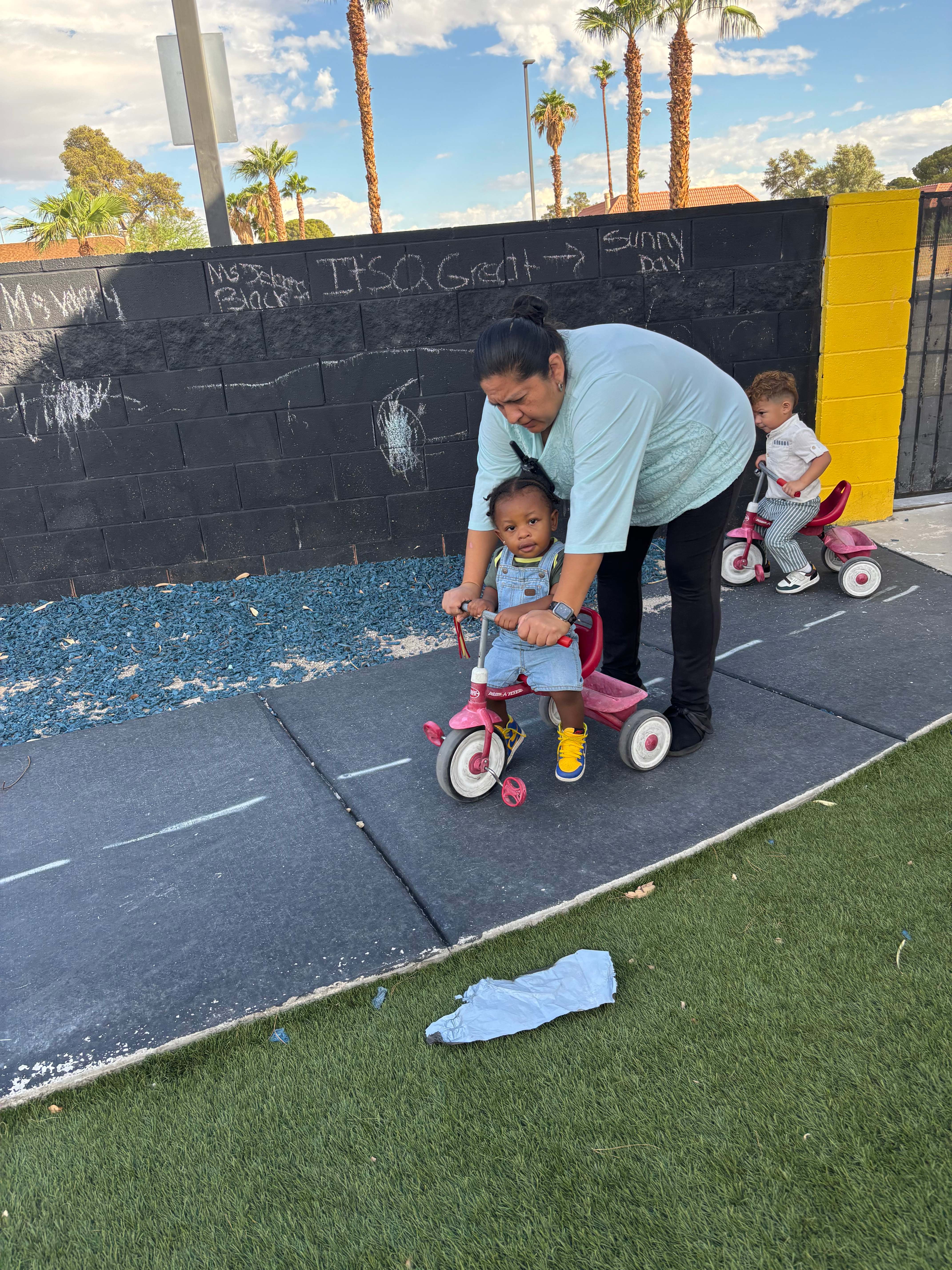 A caring Sunny Day Foundation mentor helping children ride tricycles along the outdoor track in front of the chalk wall saying It is a Great Sunny Day