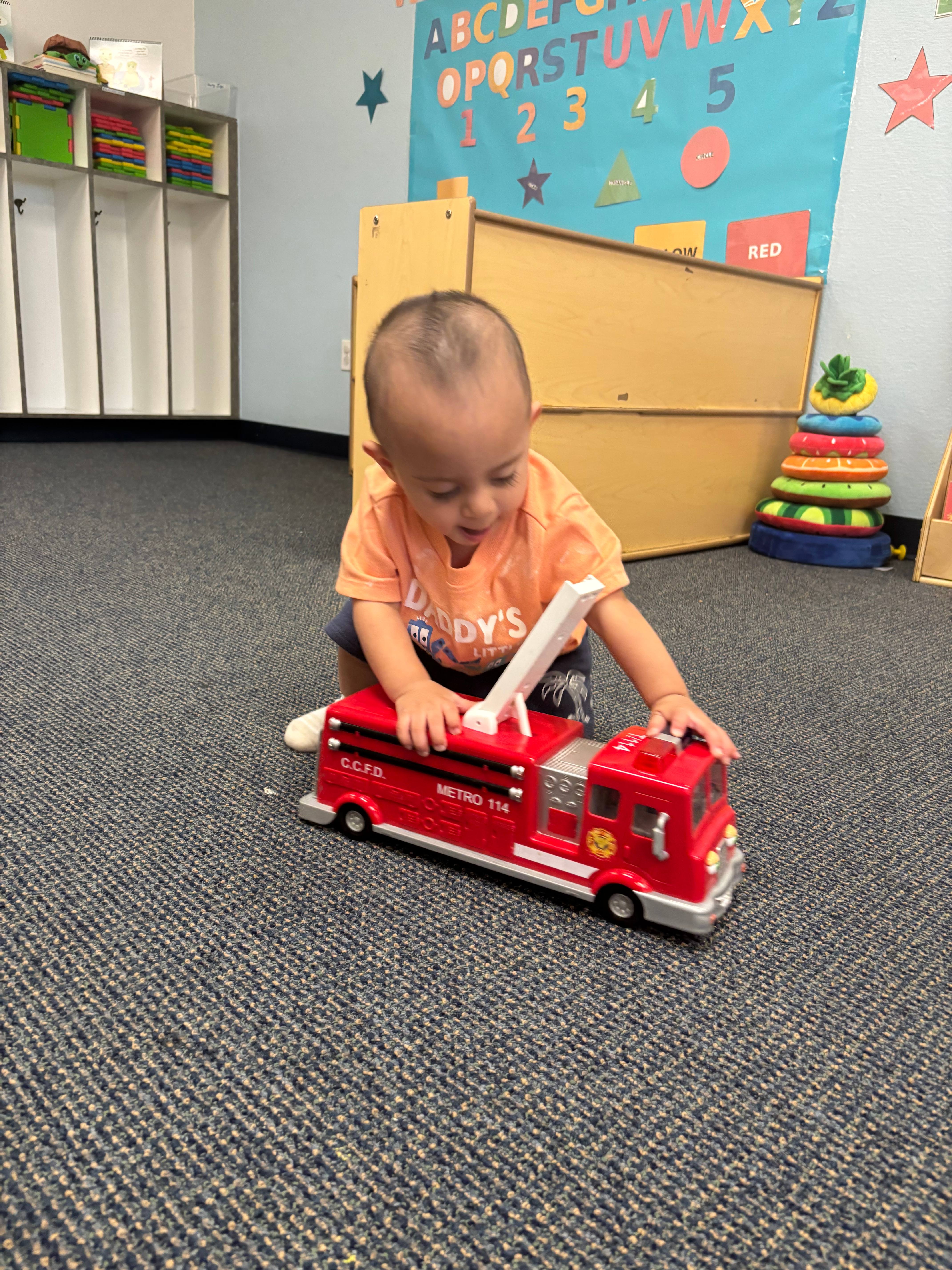 Young child exploring a red toy fire truck in an early learning playroom with educational charts