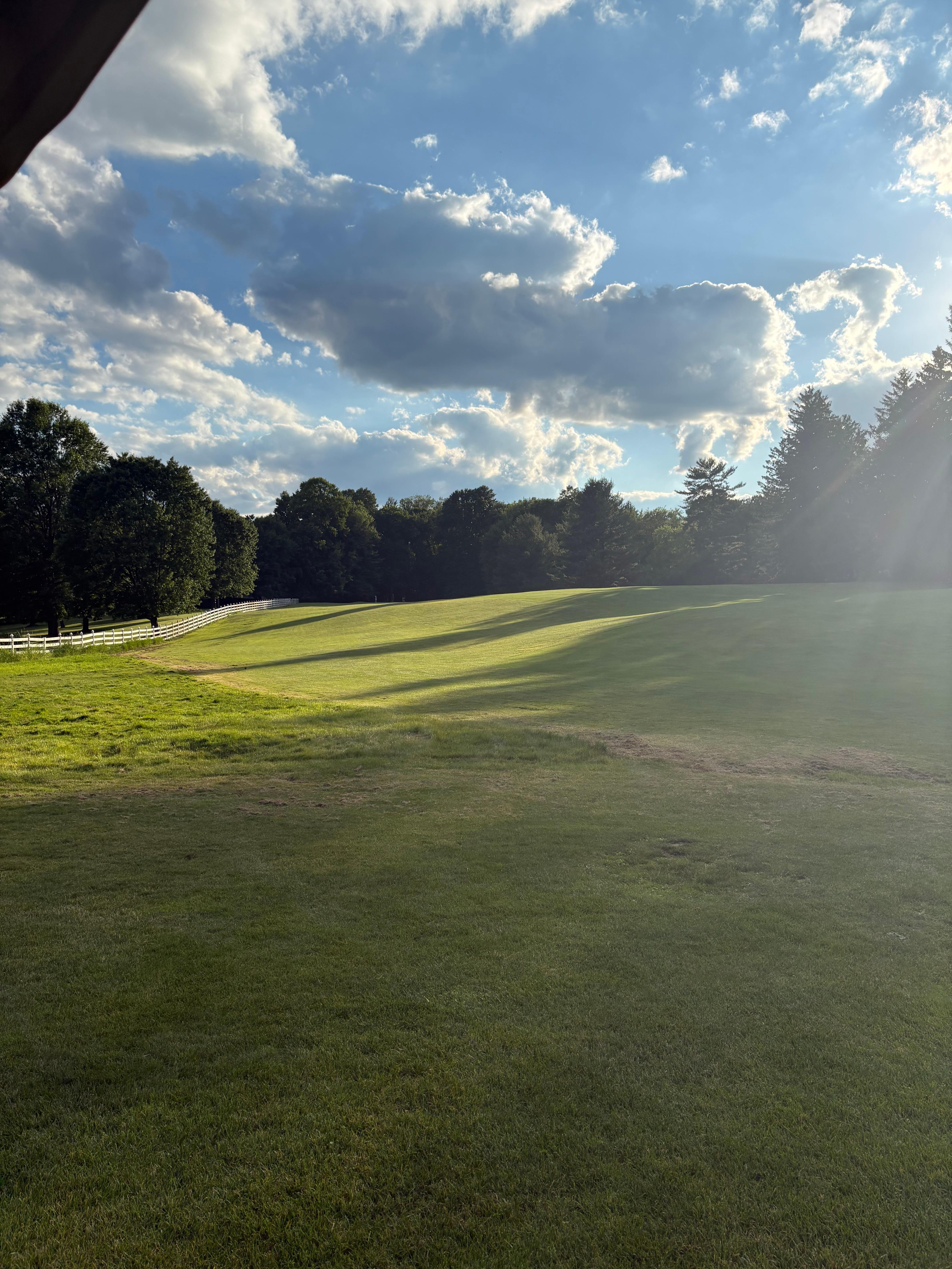 Sunlit fairway with a white rail fence and dramatic clouds