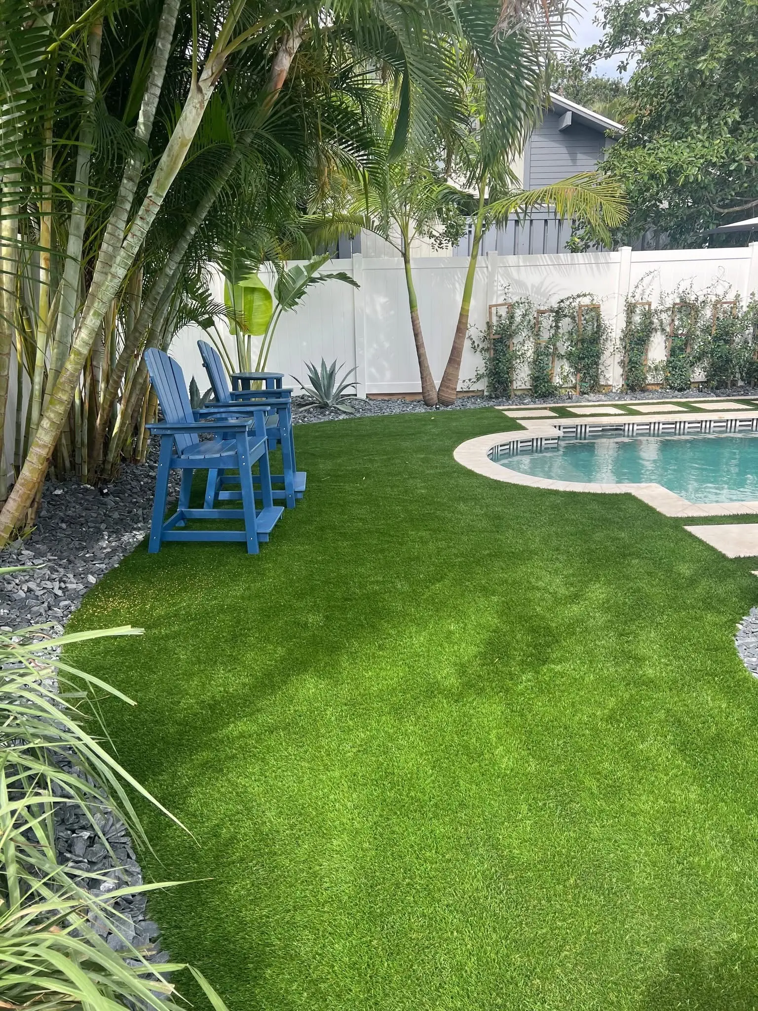 Artificial turf and landscape installation by Integrity Landscape showing a lush, emerald-green synthetic lawn surrounding a freeform pool with travertine coping, two blue Adirondack chairs, areca palms, and a white vinyl privacy fence in a Jupiter backyard